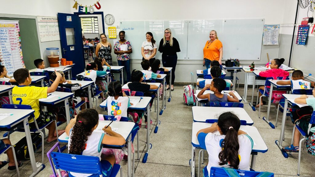 Esta imagem mostra uma sala de aula de ensino fundamental em pleno funcionamento. Ambiente: A sala está organizada com carteiras individuais em fileiras, onde os alunos estão sentados de costas para a câmera, concentrados em tarefas. Há cartazes nas paredes com listas de nomes ("Chamadinha"), uma lousa branca grande ao fundo e decorações, incluindo letras penduradas no alto da porta. Pessoas: No centro da sala, de frente para os alunos, há um grupo de cinco mulheres adultas, possivelmente professoras, coordenadoras ou convidadas, observando a turma. Elas estão em pé, posicionadas entre as fileiras de carteiras e a lousa. Alunos: A maioria das crianças veste uniformes escolares, muitas com camisas que têm a palavra "Educa" estampada nas costas. Um aluno à esquerda usa uma camisa amarela com o nome "RONALDO" e o número "7" nas costas. Outra aluna, à direita, está vestindo uma blusa rosa brilhante. Clima: O ambiente parece ser de uma atividade escolar regular ou talvez uma visita pedagógica, com um clima organizado e colaborativo. Há materiais escolares e mochilas espalhados pelas mesas e pelo chão.