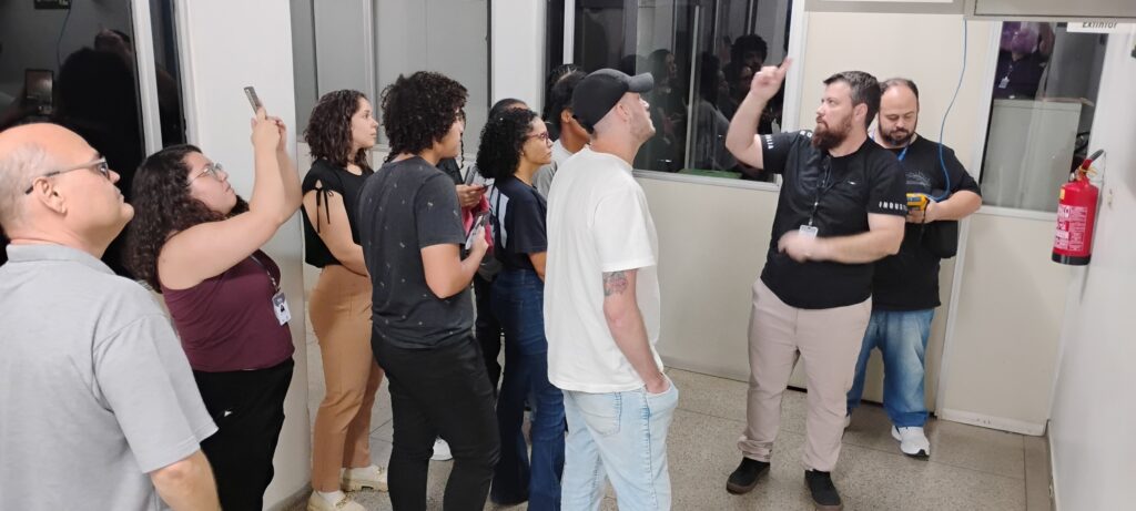 A cena ocorre em um corredor de escritório, possivelmente em uma visita técnica ou demonstração. Há um grupo de pessoas em pé, interagindo com um homem que parece estar explicando algo. A iluminação é artificial, provavelmente de fluorescentes, criando um ambiente claro, mas sem sombras muito marcadas. No centro da imagem, um homem com barba, vestindo uma camiseta preta com um emblema no peito e calças bege claras, gesticula com o braço direito, apontando para cima, como se estivesse indicando um ponto ou explicando uma instrução. Ele está virado de lado, parcialmente em direção a outro homem. À sua esquerda, um homem com boné preto e camiseta branca está olhando para onde o outro homem aponta. Ele tem um distintivo de tatuagem em seu braço esquerdo visível. Ele tem as mãos nos bolsos da calça jeans clara que veste. Um pouco mais à esquerda, uma mulher com cabelos cacheados escuros, vestindo uma camiseta vinho, olha para o celular que segura em suas mãos. Ao seu lado, um homem mais velho com óculos e careca, vestindo uma camiseta cinza clara, também levanta o olhar na mesma direção. Atrás do homem com a camiseta preta e a mulher com o celular, há outras pessoas, a maioria mulheres, com cabelos cacheados e escuros, vestindo roupas casuais. Algumas parecem estar prestando atenção, enquanto outras estão mais ao fundo e menos visíveis. Um homem com uma camiseta preta está mais ao fundo, olhando para um dispositivo amarelo em suas mãos, que pode ser um medidor ou ferramenta. No fundo, há portas, paredes claras e divisórias de vidro. Há também um extintor de incêndio vermelho montado na parede, próximo a uma porta com a palavra "EXTINTOR" escrita acima. A presença do extintor e do homem com o dispositivo sugere um ambiente de trabalho técnico ou industrial. As cores predominantes são tons neutros: bege, branco, cinza e preto, com alguns toques de cor nas roupas das pessoas e no extintor. A composição é dividida entre o grupo de pessoas em primeiro plano e o fundo do corredor, com o homem que gesticula servindo como ponto focal. A imagem parece ter sido tirada em um ângulo ligeiramente para cima.
