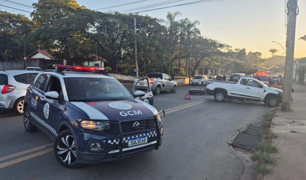 A imagem mostra uma cena de ocorrência de trânsito em uma via pública, com a presença da Guarda Civil Municipal (GCM) de Piracicaba. Aqui estão os detalhes observados: Veículo Oficial: Em primeiro plano, há uma viatura da GCM (um Volkswagen T-Cross caracterizado) com o giroflex ligado e posicionado para bloquear ou sinalizar parte da via. Ocorrência: No centro da imagem, é possível ver uma motocicleta caída no chão, próxima a um triângulo de sinalização, indicando um acidente. Outros veículos: Logo atrás da moto, há uma picape branca (Fiat Strada) parada, possivelmente envolvida ou parada para prestar auxílio, além de outros carros circulando ou parados ao fundo. Ambiente: A cena ocorre em um cruzamento urbano durante o dia. Há uma equipe de pessoas (possivelmente agentes ou populares) observando perto da picape e outra viatura, também com luzes de emergência, visível mais ao fundo.
