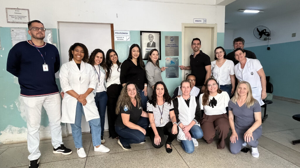 Uma fotografia de grupo de uma equipe profissional, composta por diversas pessoas, posa alegremente em um ambiente de hospital ou clínica, celebrando um momento de conquista ou comemoração de trabalho. A composição da cena é organizada com os membros da equipe dispostos em duas fileiras, uma em pé e outra agachada, em frente a uma parede de hospital onde há placas comemorativas. O enquadramento é amplo, capturando todo o grupo e parte do ambiente clínico. A interação entre as pessoas é positiva, com muitos sorrindo para a câmera, enquanto alguns apontam para a placa fixada na parede, indicando um marco institucional ou uma homenagem recente. A disposição equilibra o peso visual dos integrantes, criando uma imagem coesa e harmoniosa. Os sujeitos, um grupo diversificado de homens e mulheres adultos, vestem uma mistura de trajes profissionais, incluindo jalecos brancos, roupas casuais de escritório e uniformes de saúde. Suas expressões são variadas entre sorrisos genuínos e olhares atentos, transmitindo uma sensação de camaradagem e orgulho profissional. As poses são naturais, com mãos cruzadas ou apoiadas em colegas, reforçando a conexão interpessoal entre os membros da equipe. Cada indivíduo contribui para a atmosfera de trabalho colaborativo, com características físicas variadas que refletem a diversidade do corpo clínico presente. Esta imagem é uma fotografia digital de registro, com estilo documental típico de fotos de grupo em ambientes corporativos ou institucionais. A execução técnica é direta, focada em capturar a nitidez de todos os integrantes, mantendo um equilíbrio de cores que reflete a luz interna do espaço. Não há filtros estilísticos complexos, sendo uma representação fiel de um momento cotidiano, com foco na clareza e na identidade dos sujeitos e do local, cumprindo seu propósito de ser um registro memorial daquela equipe específica. O cenário é um corredor ou área de espera de uma unidade de saúde, caracterizado pelas paredes pintadas em tons suaves de azul e branco. A iluminação é difusa, vinda de fontes artificiais no teto, que iluminam uniformemente o grupo e o ambiente. Elementos como placas informativas, uma moldura com uma foto em preto e branco e um ventilador na parede superior reforçam a natureza institucional do local. A atmosfera geral é profissional e acolhedora, transmitindo uma sensação de estabilidade, dedicação e união entre os colegas de trabalho que ali se encontram.