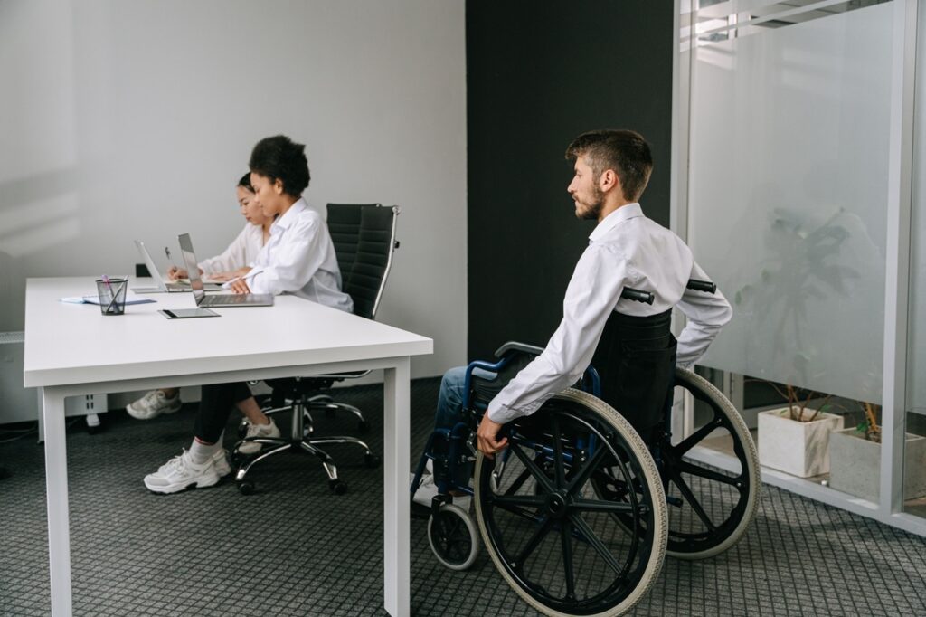 A imagem mostra um ambiente de escritório moderno e acessível. Em primeiro plano, um homem sentado em uma cadeira de rodas se aproxima de uma mesa branca, onde duas colegas trabalham usando um notebook e outros materiais. O homem veste camisa branca e usa um suporte preto no tronco. Ao fundo, há cadeiras de escritório, paredes claras, divisórias de vidro e uma planta. A cena destaca inclusão e acessibilidade no ambiente profissional.