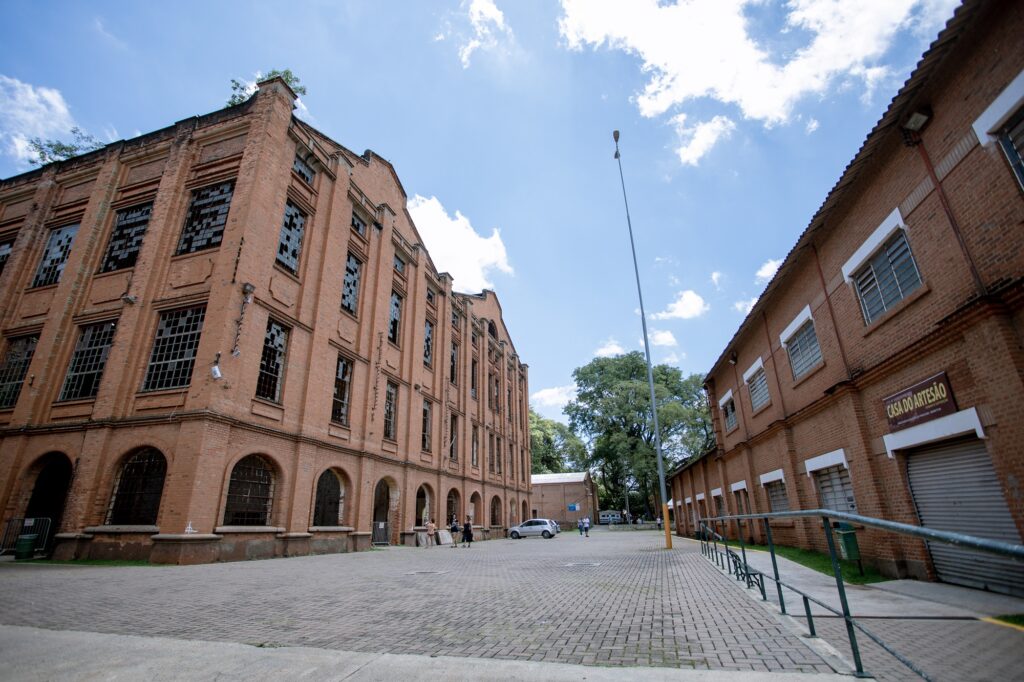 A imagem mostra um complexo de edifícios industriais históricos construídos em tijolos à vista sob um céu azul ensolarado com poucas nuvens. Edifício à esquerda: Uma grande estrutura imponente de vários andares, em estilo arquitetônico industrial antigo. Suas janelas altas possuem arcos e quadros de ferro, sendo que muitas estão quebradas ou desprovidas de vidros. No topo e nas cornijas da fachada, há pequenas vegetações crescendo. Edifício à direita: Uma construção mais baixa, também em tijolos aparentes, mais bem conservada. Há também uma rampa/escada com corrimão de metal verde ao longo deste prédio. Espaço central: Uma ampla praça ou pátio pavimentado com paralelepípedos e blocos intertravados. Ao centro, vê-se um poste alto de iluminação, alguns carros estacionados ao fundo e poucas pessoas caminhando pelo local. Ao fundo, sobressaem árvores de grande porte.