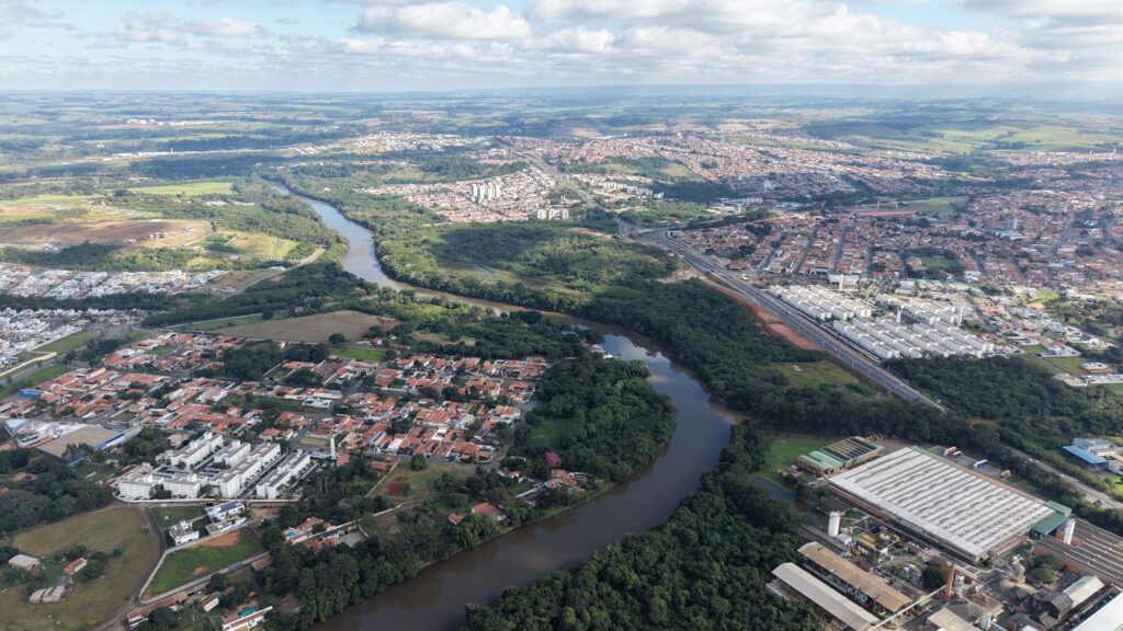 Esta é uma vista aérea impressionante de uma região que mistura áreas urbanas e natureza. Aqui estão os pontos principais: O Rio: O elemento central que domina a paisagem é um rio sinuoso que corta a cena de cima para baixo, rodeado por uma densa vegetação nativa nas suas margens, que forma um corredor verde bem preservado. Áreas Urbanas: De ambos os lados do rio, nota-se o crescimento urbano. À esquerda, vemos um bairro residencial com casas de telhados claros e alguns conjuntos de prédios ou galpões. À direita, há uma área mais densa, com bairros residenciais, uma rodovia movimentada que corta a cidade diagonalmente e, no canto inferior direito, um grande complexo industrial com galpões de telhados metálicos brancos. Geografia: A paisagem ao fundo se estende até o horizonte, mostrando uma área extensa de campos, possivelmente plantações ou áreas rurais, sob um céu com nuvens espalhadas. Sensação Geral: É uma imagem que retrata bem o contraste entre o desenvolvimento humano (casas, estradas, indústrias) e a preservação ambiental ao longo dos cursos d'água.