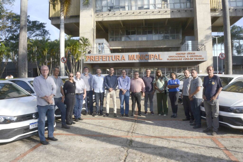 A imagem mostra um grupo numeroso de pessoas reunido em frente ao Centro Cívico Educacional e Cultural Florivaldo Coelho Prates, identificado na fachada. O prédio tem arquitetura ampla, com colunas, grandes janelas, grades metálicas e palmeiras na entrada. Na parte inferior, várias pessoas posam para uma foto ao lado de um carro branco. A maioria usa camisas em tons claros, e algumas vestem crachás ou camisetas com símbolos. A cena acontece ao ar livre, em um dia ensolarado.