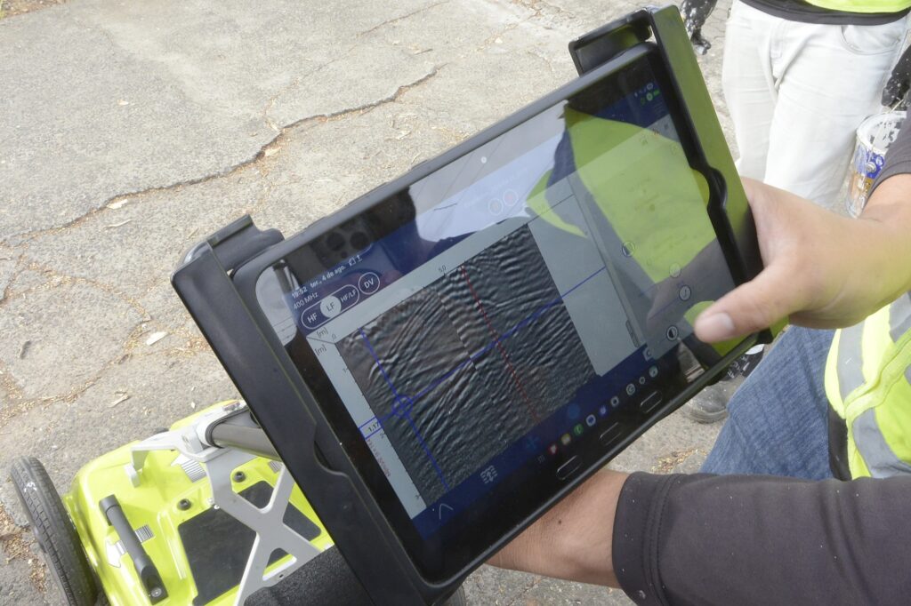 Na imagem vemos uma cena de trabalho de campo com um equipamento portátil de inspeção: Em primeiro plano, as mãos de uma pessoa seguram um tablet dentro de uma capa preta; a tela mostra um gráfico de varredura (linhas e padrões ondulados) — provavelmente leitura de um equipamento geofísico ou radar de solo (GPR). A pessoa veste colete de alta visibilidade amarelo e calça jeans; só parte do braço e do torso aparecem — nenhuma face é identificada. Abaixo e à esquerda do tablet há uma unidade verde com rodas e estrutura metálica, parecendo ser o carrinho/antena do equipamento que varre o solo. O chão é asfalto rachado; há folhas secas espalhadas e iluminação de dia claro. A composição sugere que a equipe está realizando medição ou mapeamento do subsolo em via pública.