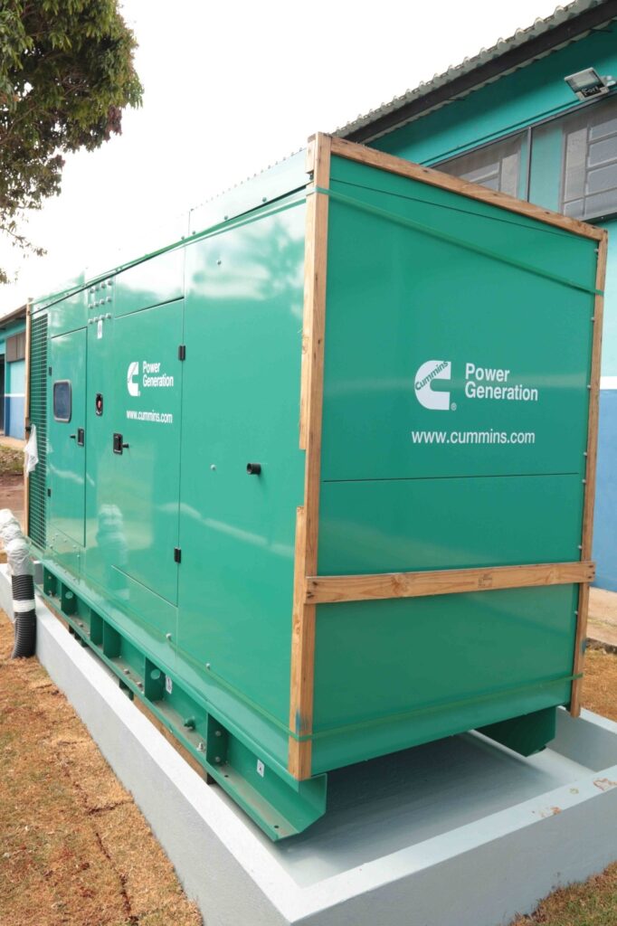 A imagem mostra um grande gerador elétrico industrial verde, instalado sobre uma base de concreto ao lado de um edifício. O equipamento tem a marca Cummins Power Generation e o endereço www.cummins.com estampados na carcaça. Ele possui portas de acesso, grades de ventilação, painéis metálicos e uma tubulação flexível na lateral. Uma estrutura de madeira protege parte do gerador. Ao fundo, há uma construção pintada de azul-turquesa, uma árvore e um gramado seco.