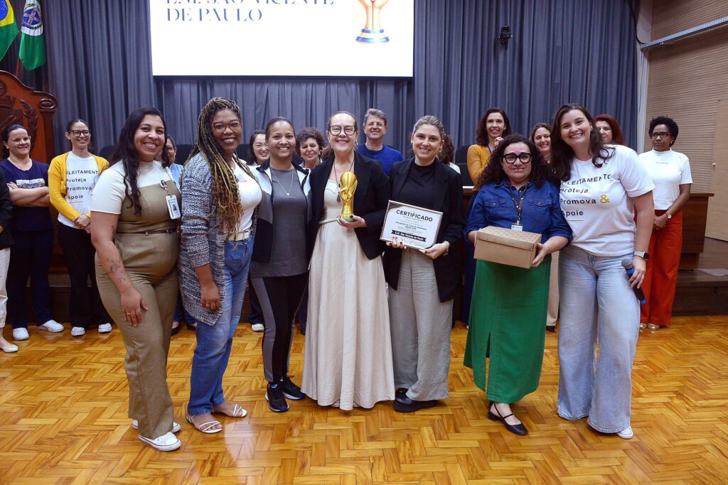 A imagem mostra um grupo de pessoas em um ambiente que parece ser um auditório, celebrando uma conquista relacionada à "Copa do Aleitamento". Aqui estão os detalhes principais: O Evento: É uma premiação. No centro, uma pessoa segura um troféu dourado que lembra a taça da Copa do Mundo, e outra pessoa segura um certificado escrito "Vencedores da Copa do Aleitamento, edição 2026", especificando "E.M. São Vicente de Paulo". As Pessoas: Há um grupo grande, majoritariamente feminino, vestindo roupas casuais e profissionais. Muitas usam camisetas com a frase "Aleitamento: Proteja, Promova & Apoie". Elas estão sorridentes, posando para uma foto de comemoração. O Cenário: Elas estão em um palco ou frente de auditório, com piso de madeira, cortinas cinzas ao fundo e uma tela de projeção onde se lê parte do nome da escola "E.M. São Vicente de Paulo". Detalhes Adicionais: Algumas pessoas seguram microfones ou caixas de presente, indicando que a celebração envolveu discursos ou entrega de lembranças. O clima é de união e orgulho pela conquista.