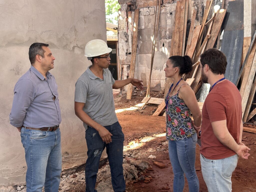 Na imagem, quatro pessoas estão em um canteiro de obras, cercadas por paredes inacabadas, tábuas de madeira e materiais de construção espalhados. Um homem de capacete branco e camisa polo cinza parece conduzir a conversa, apontando ou explicando algo aos demais. Os outros três, vestidos de forma casual — dois de jeans e um com blusa florida — escutam atentamente, sugerindo uma reunião de inspeção ou planejamento sobre o andamento da obra.