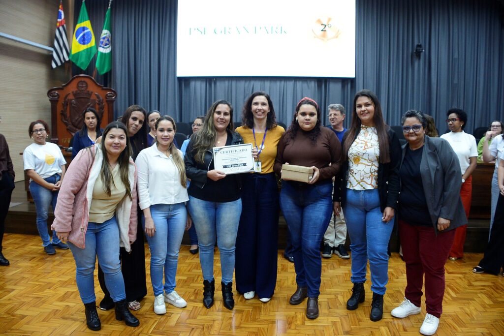A imagem mostra um grupo de pessoas em um auditório, celebrando uma premiação na "Copa do Aleitamento, edição 2026". Aqui estão os detalhes principais: A Celebração: No centro, uma das participantes segura um certificado que identifica a equipe vencedora como "USF Gran Park", com a inscrição "2º" visível no telão ao fundo. Outra participante segura uma caixa, que parece ser um presente ou troféu da premiação. O Grupo: É um grupo majoritariamente feminino, posando sorridente para a foto. Elas vestem roupas casuais e profissionais (jeans, blusas, jaquetas). O Ambiente: Elas estão em um auditório com piso de madeira, cortinas cinzas ao fundo e bandeiras (do Brasil e do Estado de São Paulo) na lateral esquerda. Contexto: A atmosfera é de comemoração e reconhecimento de uma equipe de saúde (provavelmente uma Unidade de Saúde da Família - USF) por seu trabalho ou desempenho na "Copa do Aleitamento".
