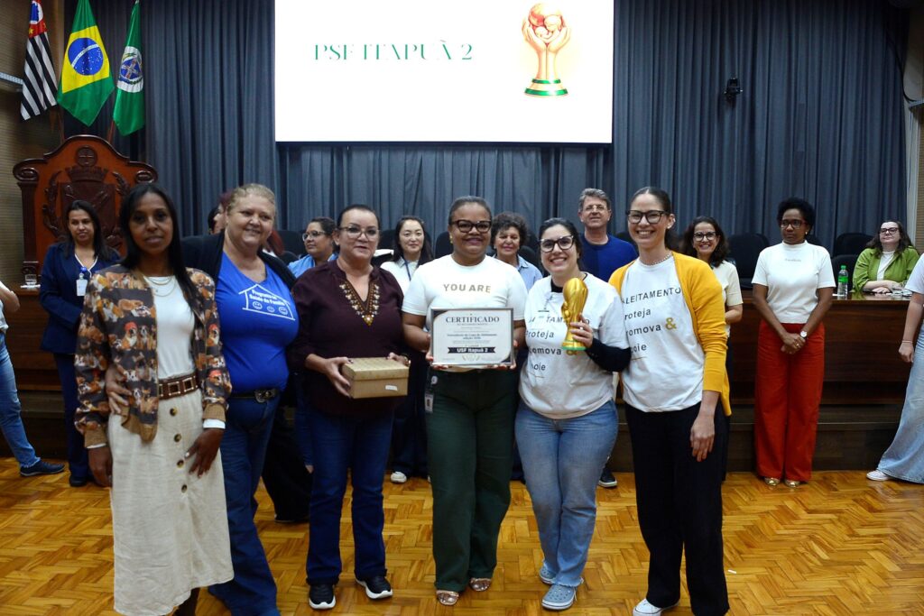 A imagem mostra um grupo de pessoas em um auditório, celebrando uma premiação na "Copa do Aleitamento, edição 2026". Aqui estão os detalhes principais: A Celebração: No centro, uma das participantes segura um certificado que identifica a equipe vencedora como "USF Itapuã 2", com o mesmo nome também exibido em destaque no telão ao fundo. Ao lado, outra participante segura um troféu dourado. O Grupo: É um grupo de profissionais (majoritariamente feminino), posando sorridente para a foto. Elas vestem roupas casuais e algumas usam camisetas com a frase "Aleitamento: Proteja, Promova & Apoie". O Ambiente: Estão em um auditório com piso de madeira, cortinas cinzas ao fundo e bandeiras (do Brasil e do Estado de São Paulo) na lateral esquerda. Contexto: A atmosfera é de comemoração pelo reconhecimento do trabalho da unidade de saúde (USF Itapuã 2) na promoção do aleitamento materno.