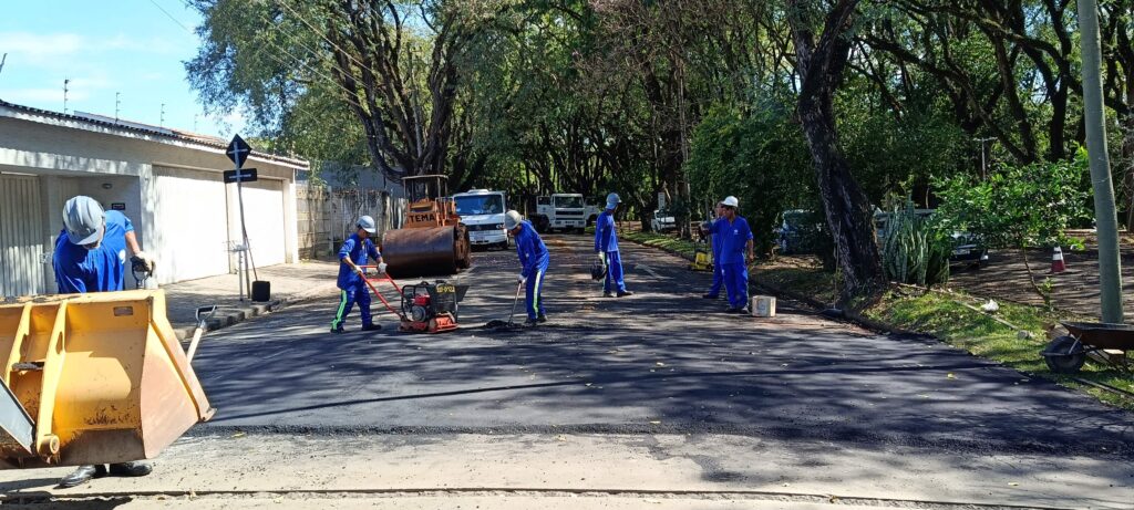 Nesta imagem, vemos uma equipe de trabalhadores realizando obras de pavimentação em uma rua arborizada. Aqui estão os detalhes principais: A Ação: Vários homens vestindo uniformes azuis e capacetes brancos estão trabalhando ativamente no asfalto fresco, que ainda está bem escuro. Um deles opera uma compactadora de solo (sapinho), outro parece espalhar material com uma pá, e há um rolo compactador da marca "TEMA" parado logo atrás. O Ambiente: É uma rua residencial tranquila, cercada por árvores grandes que criam uma sombra densa sobre a via. À esquerda, há uma casa com portão branco e muros altos. Equipamento: Além do maquinário pesado (rolo compactador e caminhões ao fundo), há uma caçamba de uma máquina (possivelmente um trator ou retroescavadeira) em primeiro plano à esquerda. Clima: O dia parece ensolarado e claro, com o sol projetando sombras nítidas das árvores sobre o novo asfalto.