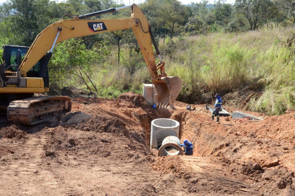 Na imagem, vemos um canteiro de obras onde uma escavadeira amarela da Caterpillar abre uma vala em meio ao solo, cercado por gramado e árvores. Dentro da trincheira, estão sendo posicionados tubos de concreto para instalação de sistemas de drenagem ou esgoto. Dois trabalhadores com uniformes azuis e equipamentos de proteção participam da atividade: um dentro da vala, ajustando os tubos, e outro na borda, supervisionando o processo. O braço da escavadeira auxilia na movimentação das peças pesadas, evidenciando a integração entre força mecânica e trabalho humano em uma etapa essencial da engenharia civil voltada à infraestrutura subterrânea.