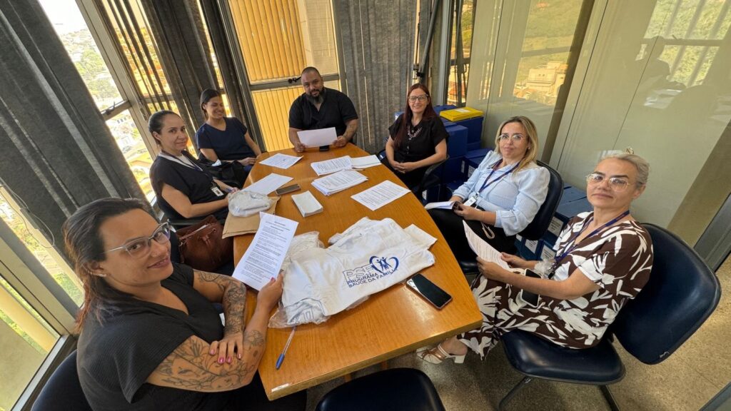 A imagem mostra um grupo de sete pessoas reunido ao redor de uma mesa de madeira, em uma sala com janelas amplas e persianas verticais. Algumas pessoas seguram folhas de papel, enquanto outras observam os documentos sobre a mesa. Sobre a mesa também há celulares, canetas, uma bolsa, sacolas plásticas e camisetas brancas com o logotipo “Programa Saúde da Família”. Os participantes usam crachás e parecem estar envolvidos em uma atividade ou reunião de trabalho. Ao fundo, pelas janelas, é possível ver parte da paisagem urbana e áreas verdes.