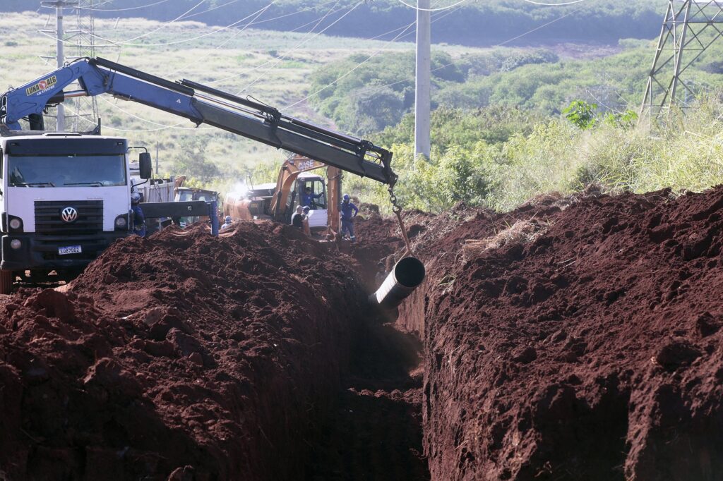 A cena retratada é uma obra de construção em uma área rural ou em desenvolvimento, onde um canal profundo foi escavado no solo avermelhado. Vários objetos e pessoas estão envolvidos no processo de instalação de um grande tubo dentro deste canal. No lado esquerdo da imagem, domina um caminhão branco com detalhes em preto e o logo da Volkswagen na grade frontal. Um braço hidráulico azul claro, com a inscrição "LUNA ALG LN 4757BR", está acoplado ao caminhão e estende-se por cima da cena. Este braço está a segurar um tubo cinzento escuro, com anéis prateados visíveis, que está a ser baixado para dentro do canal. Dentro do canal, no centro da imagem, uma escavadora amarela, com a marca "CASE" visível, está posicionada. Várias pessoas vestindo uniformes de trabalho azuis e capacetes brancos estão presentes. Algumas parecem estar a coordenar a descida do tubo, enquanto outras operam a maquinaria. Há montes de terra avermelhada solta em ambos os lados do canal, indicando que a escavação é recente. O fundo da imagem mostra uma paisagem natural com vegetação verde densa e colinas. Uma torre de alta tensão com fios elétricos suspensos atravessa o céu, sugerindo a proximidade de infraestruturas. O céu é claro e azul, com alguns raios de sol a iluminar a cena, criando sombras distintas no solo. A composição da imagem é dominada pela diagonal acentuada do canal escavado e pelo braço hidráulico. A terra avermelhada cria um contraste forte com a vegetação verde e o azul do céu e do braço hidráulico. Os detalhes de textura do solo, as ferramentas e as vestimentas dos trabalhadores adicionam realismo à cena, que retrata um momento de trabalho intensivo e coordenação em um projeto de infraestrutura. As cores predominantes são o vermelho-acastanhado da terra, o verde da vegetação, o branco e o azul dos veículos e dos trabalhadores, e o cinzento dos tubos.