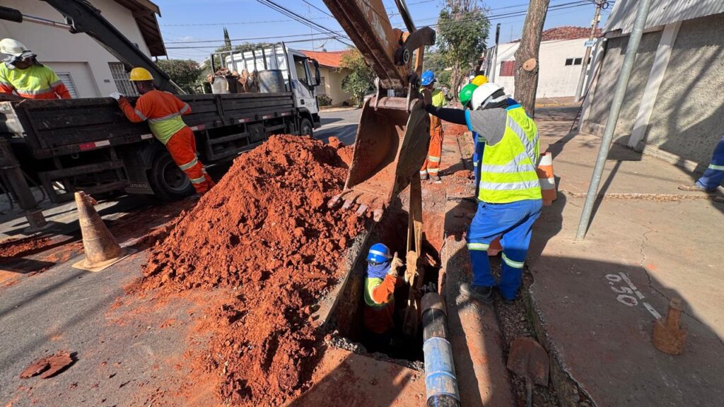 A cena retrata uma obra pública em uma rua residencial, onde trabalhadores estão escavando para instalar ou reparar tubulações subterrâneas. A imagem é dominada por tons terrosos de laranja e marrom, provenientes da terra revolvida, contrastando com as cores vibrantes dos uniformes de segurança dos trabalhadores e o azul do céu. No centro da imagem, uma retroescavadeira, com sua caçamba cheia de terra avermelhada, está posicionada sobre uma vala recém-escavada. A caçamba, de cor laranja desbotado, exibe marcas de uso e terra incrustada, sugerindo trabalho árduo. A haste metálica da retroescavadeira se projeta diagonalmente para cima, adicionando dinamismo à composição. Um trabalhador, vestindo um colete de segurança amarelo fluorescente sobre um macacão azul e um capacete branco, está em pé, próximo à retroescavadeira, observando a operação. Ele está de costas para o espectador, com a mão direita ligeiramente erguida, como se estivesse dando uma instrução ou verificando algo. Seus sapatos de segurança pretos e um detalhe verde em sua perna complementam seu traje. Dentro da vala, um outro trabalhador, com um capacete azul, está agachado, trabalhando em meio aos canos recém-instalados. Ele veste um macacão laranja e amarelo e parece estar manipulando a tubulação. À esquerda da cena, um caminhão de carga, com a parte traseira preta e um guindaste parcialmente visível, está estacionado. Dois trabalhadores com uniformes laranja e amarelo e capacetes brancos estão próximos ao caminhão, um deles inclinado sobre a lateral, como se estivesse supervisionando o carregamento ou descarregamento de materiais. Um cone de trânsito laranja e branco está posicionado no asfalto, perto do caminhão e da terra acumulada. O solo é uma mistura de asfalto rachado e terra solta, com pedaços de terra espalhados pela superfície. Há tubos azuis e pretos visíveis na vala, indicando a infraestrutura sendo trabalhada. Ao fundo, casas com telhados vermelhos e muros brancos compõem o cenário urbano. Árvores com folhagem verde e troncos claros pontuam o ambiente, e um poste de luz com fios elétricos atravessa a parte superior direita da imagem. A composição é dinâmica, com as linhas diagonais da retroescavadeira e da vala guiando o olhar do espectador através da ação. As sombras projetadas indicam que a foto foi tirada em um dia ensolarado, com a luz incidindo de um ângulo ligeiramente alto. A textura da terra, os detalhes dos uniformes de alta visibilidade e as marcas de uso nos equipamentos transmitem uma sensação de autenticidade e trabalho manual. Há uma marca branca no asfalto com o número "0.50", possivelmente indicando uma medida ou profundidade.