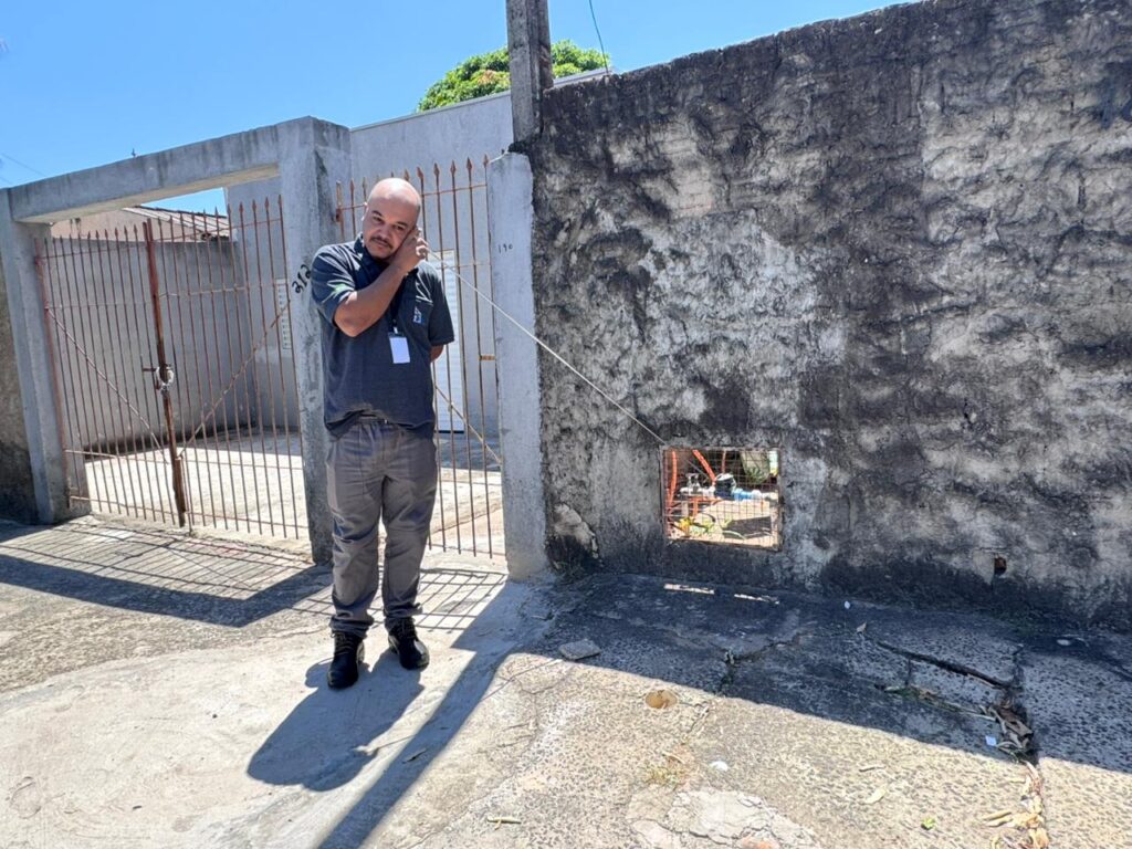 Um homem careca, usando uma camiseta cinza com um crachá, calça cinza e botas pretas, está em pé em uma rua de concreto, falando ao telefone. Ele está perto de um portão de metal enferrujado que leva a uma casa. À direita, há um muro de pedra irregular com uma abertura quadrada, parcialmente visível, revelando um espaço interior com objetos e equipamentos. O céu está claro e azul, e as sombras projetadas pelo sol indicam que é um dia ensolarado. As cores predominantes são os tons de cinza do concreto e da parede, o marrom enferrujado do portão e o azul do céu. A composição coloca o homem em primeiro plano, com os elementos arquitetônicos servindo de pano de fundo e detalhe.