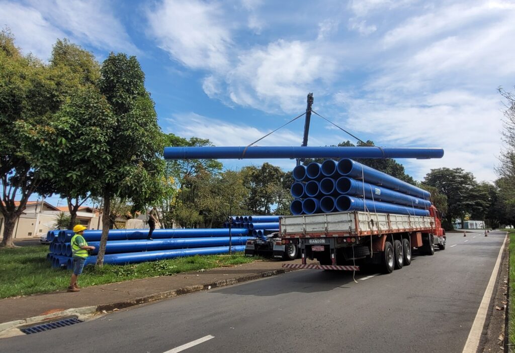 A cena retrata um local ao ar livre, provavelmente uma rua com um canteiro de obras ou área de armazenamento. O foco principal são os tubos azuis de grande diâmetro. Um caminhão carregado com uma pilha desses tubos está posicionado à direita da imagem. O caminhão é de cor laranja com uma carroceria branca e detalhes em madeira, e tem uma placa visível com os dizeres "RANDON" e um número de placa. Vários eixos com rodas duplas são visíveis na parte traseira do caminhão. Uma grua, presa ao caminhão, está erguendo um único tubo azul longo horizontalmente, no centro da imagem, acima da carga empilhada. À esquerda, uma grande quantidade de tubos azuis está empilhada no chão, ao lado de uma calçada. Uma pessoa vestindo um capacete amarelo de segurança, um colete refletivo verde e shorts azuis está em pé perto desses tubos, olhando para eles. Há também uma segunda pessoa em pé sobre os tubos empilhados no caminhão, parecendo supervisionar a operação. O fundo da cena é composto por árvores frondosas e verdes em tons variados, algumas com folhagens mais densas e outras mais esparsas. Uma casa com telhado e paredes claras, em tons de bege e branco, pode ser vista entre as árvores à esquerda. A rua asfaltada em primeiro plano tem faixas brancas e uma sarjeta com uma grade de drenagem. O céu é de um azul vibrante, pontuado por nuvens brancas e finas que se espalham, criando um contraste agradável com o azul dos tubos e o verde da vegetação. A iluminação geral sugere um dia claro e ensolarado. A composição da imagem coloca o caminhão e a carga de tubos como elementos centrais, com a grua adicionando um elemento de ação e dinâmica. A presença das pessoas indica uma atividade em andamento. As cores predominantes são o azul dos tubos, o verde das árvores e o azul do céu, com o laranja do caminhão servindo como um ponto de destaque. A linha diagonal criada pela rua e a perspectiva do caminhão guiam o olhar do espectador através da cena.