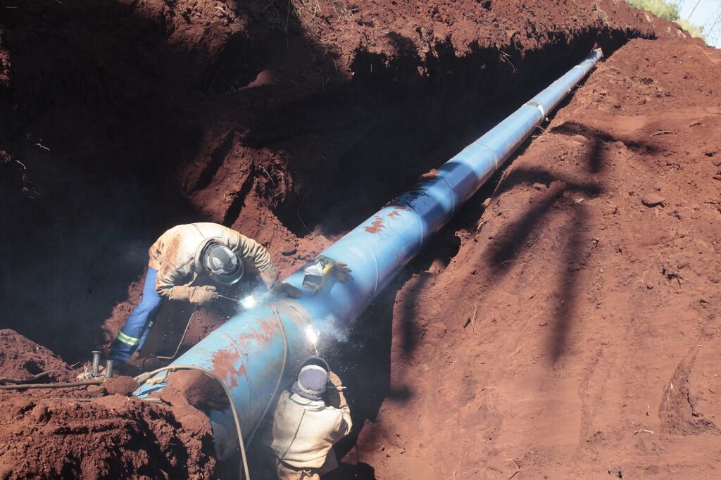 Uma imagem tirada em um ângulo ligeiramente elevado mostra uma vala em terra vermelha escavada para a instalação de um longo tubo azul. O tubo, que parece ser de metal, corre diagonalmente através da vala, desaparecendo no fundo escuro da esquerda e subindo em direção ao canto superior direito. A cor vermelha da terra é proeminente, com texturas ásperas e algumas pequenas raízes e detritos visíveis nas paredes da vala. A vala é bastante profunda, criando sombras escuras em algumas áreas. Na parte inferior esquerda da imagem, dois trabalhadores estão ocupados soldando o tubo. Ambos usam equipamentos de segurança, incluindo capacetes com visores abaixados e roupas de proteção. O trabalhador mais próximo da câmera está abaixado, operando uma máquina de solda que emite uma forte luz branca e faíscas brilhantes. Ele usa um macacão azul com detalhes em verde fluorescente e luvas grossas. O outro trabalhador está um pouco mais atrás e acima, também envolvido no processo de soldagem, com suas ferramentas visíveis. A fumaça e as faíscas da soldagem criam um efeito cintilante e adicionam dinamismo à cena. O tubo azul tem um acabamento liso, mas em algumas áreas, especialmente onde está sendo soldado, pode-se ver vestígios de ferrugem ou sujeira, indicando que é um material robusto em processo de instalação. A iluminação na cena é forte, vinda de cima e ligeiramente da direita, o que realça as cores vibrantes da terra vermelha e o brilho da soldagem. A composição da imagem enfatiza a magnitude do projeto com o longo tubo serpenteando pela paisagem escavada, com os trabalhadores como elementos centrais realizando o trabalho essencial. A perspectiva cria uma sensação de profundidade e escala, mostrando o esforço envolvido na construção da infraestrutura. Há uma sombra projetada na terra no lado direito da imagem, possivelmente de uma estrutura próxima ou de parte da vala.