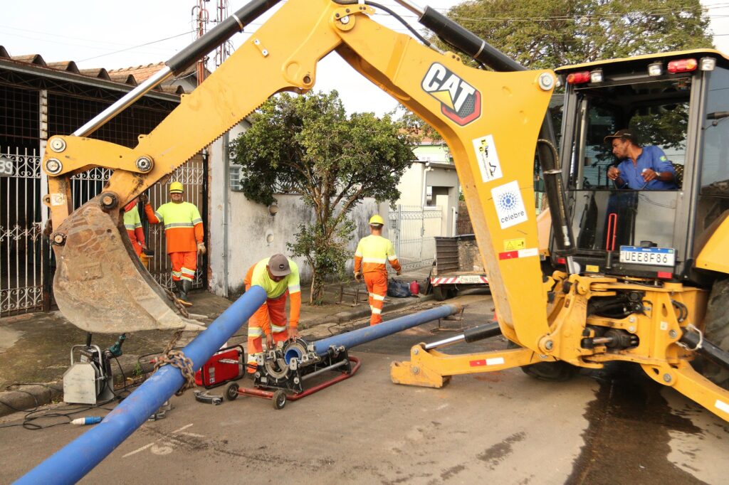 Trabalhadores realizam obra de instalação de tubulação utilizando retroescavadeira e equipamentos. A imagem mostra uma cena de obra em uma rua residencial, com foco em uma retroescavadeira amarela da marca CAT. Um homem, vestindo um capacete amarelo, colete refletivo e calças laranjas, está ao lado da máquina, supervisionando o trabalho. Outros dois trabalhadores, também com trajes de segurança refletivos em amarelo e laranja, estão envolvidos na instalação de uma grande tubulação azul que atravessa a rua. Um deles está abaixado, operando uma máquina de fusão de tubos, enquanto o outro está mais afastado, em direção à edificação ao fundo. A retroescavadeira tem sua pá carregadeira levantada, com uma corrente prendendo a tubulação. No interior da cabine da máquina, um homem com boné e camisa azul observa a cena. Vários equipamentos de construção, como uma fonte de energia portátil e a máquina de fusão, estão dispostos no chão. As casas ao redor e uma árvore com folhagem verde compõem o cenário. O clima parece ser de trabalho em andamento, com uma atmosfera funcional e dedicada. As cores predominantes são o amarelo vibrante da retroescavadeira, o azul da tubulação e o laranja e amarelo dos uniformes dos trabalhadores, contrastando com o cinza do asfalto e o branco das edificações. A composição da imagem destaca a magnitude da máquina e da tubulação, enfatizando a tarefa que está sendo executada.