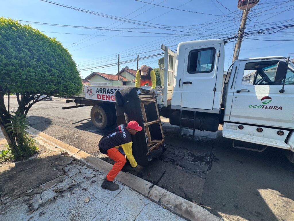 Uma fotografia documental captura dois trabalhadores municipais realizando a remoção de um móvel velho e descartado para o caminhão de coleta em um esforço comunitário de limpeza urbana sob luz solar. A composição da cena é focada em uma ação prática de trabalho, com um caminhão branco posicionado lateralmente ao longo de uma rua residencial. O ângulo da câmera, levemente elevado, abrange o veículo, a calçada e parte da via pública. A ação principal ocorre no centro, onde um trabalhador inclinado na calçada segura a base de um sofá antigo, enquanto seu colega, em cima da carroceria do caminhão, auxilia no manuseio do objeto. O enquadramento equilibra elementos humanos, mecânicos e o ambiente urbano circundante, evidenciando o processo de recolhimento de resíduos. Os dois trabalhadores estão uniformizados com camisas amarelas de mangas compridas e calças laranjas, vestindo coletes que os identificam como parte da equipe de limpeza. O indivíduo em pé na calçada usa um boné vermelho e adota uma postura curvada, exercendo esforço físico para levantar o sofá. O colega sobre o caminhão veste um boné de cor clara e atua como receptor do objeto, coordenando o carregamento. O sofá, que é o objeto central da coleta, aparece de cabeça para baixo, revelando sua estrutura de madeira desgastada e estofamento escuro, servindo como ponto focal da interação. Esta é uma fotografia digital que documenta um evento cotidiano com um estilo realista e direto. A execução técnica privilegia a clareza, utilizando uma lente grande-angular que captura tanto os indivíduos quanto a escala do caminhão e parte do ambiente. Não há manipulação artística óbvia, focando-se na representação fiel da tarefa em execução, com cores naturais e foco nítido tanto no primeiro plano, onde está o trabalhador, quanto no caminhão. O registro funciona como um retrato autêntico de serviços públicos em ação. O cenário é uma rua urbana tranquila sob uma luz do dia clara e intensa, típica de uma manhã ensolarada ou início de tarde. À esquerda, uma árvore frondosa e podada projeta uma sombra projetada sobre a calçada e o asfalto, adicionando contraste à cena. Ao fundo, percebem-se casas residenciais simples e uma rede complexa de fios elétricos cruzando o céu azul limpo. A iluminação natural é abundante, criando sombras definidas que reforçam a tridimensionalidade dos elementos e conferem um tom de atividade rotineira e utilitária, destacando o ambiente de um bairro residencial comum.