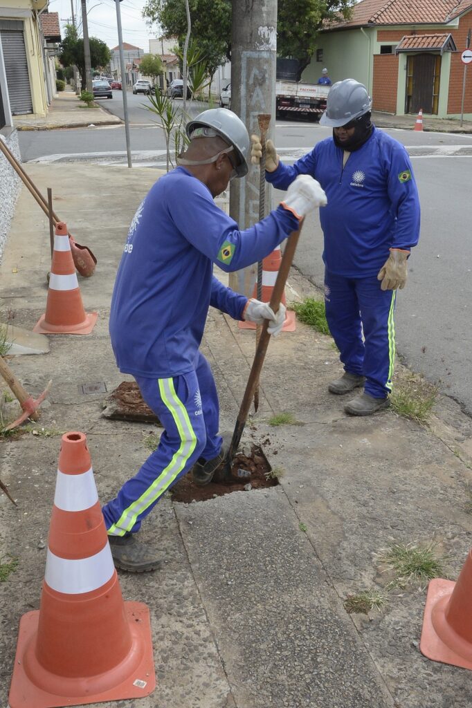 Na imagem vemos dois trabalhadores em serviço na calçada de uma rua residencial: Ambos vestem uniformes azuis, capacetes e luvas; há a bandeira do Brasil na manga de um deles e um logotipo circular no peito do uniforme. Um trabalhador está golpeando/abrindo o solo com uma barra ou pé-de-cabra, com o pé apoiado sobre a borda do buraco. O outro segura uma haste metálica (provavelmente vergalhão ou ferramenta) observando e auxiliando. Três cones de sinalização laranja delimitaram a área de trabalho na calçada/rua. No chão há um buraco no concreto, com terra exposta e fragmentos de calçada removidos. Ao fundo nota-se uma rua tranquila com casas, postes e um caminhão estacionado mais adiante. Luz do dia e pouca vegetação rasteira nas bordas da calçada.