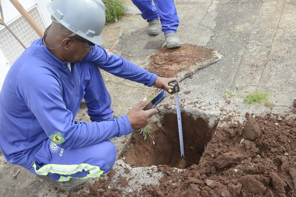 Na imagem vemos um trabalho de escavação/inspeção em uma calçada/trecho de rua: Dois trabalhadores com uniformes azuis; um está agachado perto da borda do buraco e o outro aparece em pé com apenas as pernas e botas visíveis. O trabalhador agachado usa capacete cinza, óculos de proteção e segura um celular com a mão esquerda enquanto, com a direita, segura uma trena metálica que mede a profundidade do buraco. À esquerda da roupa do operário há uma bandeira do Brasil aplicada na manga. O buraco tem bordas de concreto quebrado e solo argiloso avermelhado acumulado ao redor; pedaços de laje/remoções de calçada estão apoiados próximos à escavação. O pavimento ao redor mostra fissuras e marcas de asfalto/concreto antigo; há pequenas manchas de terra e um tufo de grama ao lado. A cena parece ser uma medição técnica ou registro fotográfico da profundidade/condição do serviço em andamento.