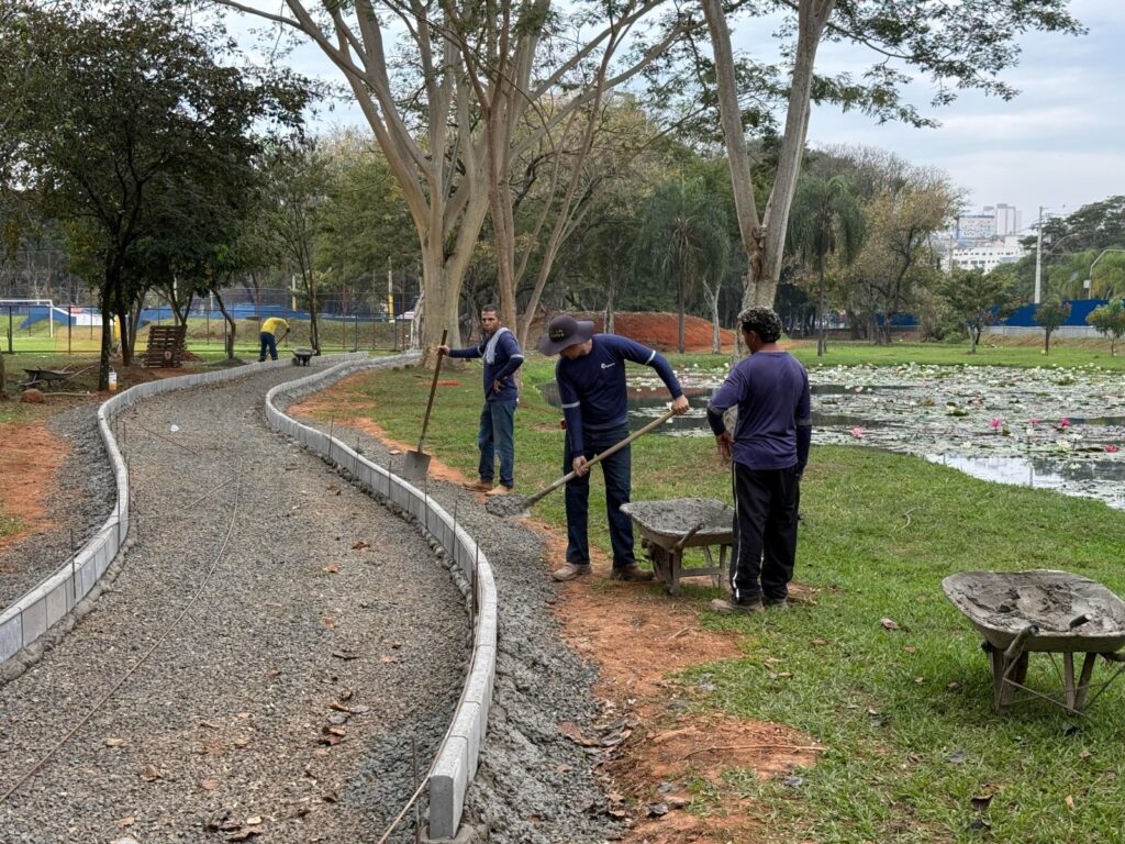A imagem mostra trabalhadores construindo ou reformando uma trilha curva em um parque. Eles espalham concreto com pás e usam carrinhos de mão cheios de material. A trilha está delimitada por bordas de concreto e coberta com pedras. À direita, há um lago com muitas plantas aquáticas e flores cor-de-rosa. Ao fundo, aparecem árvores, áreas esportivas cercadas e alguns prédios urbanos.
