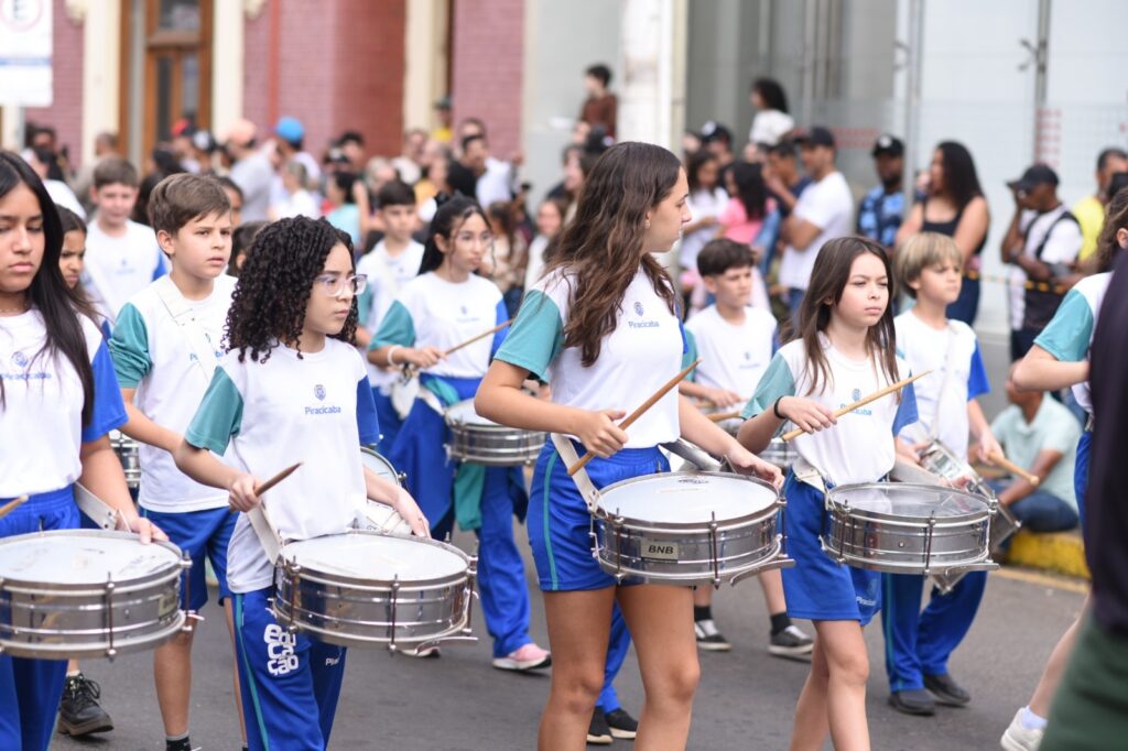 A imagem mostra um grupo de crianças e adolescentes participando de um desfile ou apresentação musical na rua. Eles vestem uniformes brancos e azuis com o nome “Piracicaba” e tocam tambores usando baquetas. Ao fundo, uma grande multidão acompanha o evento em frente a um prédio. Os participantes parecem estar organizados em uma banda marcial, caminhando e tocando instrumentos de percussão.