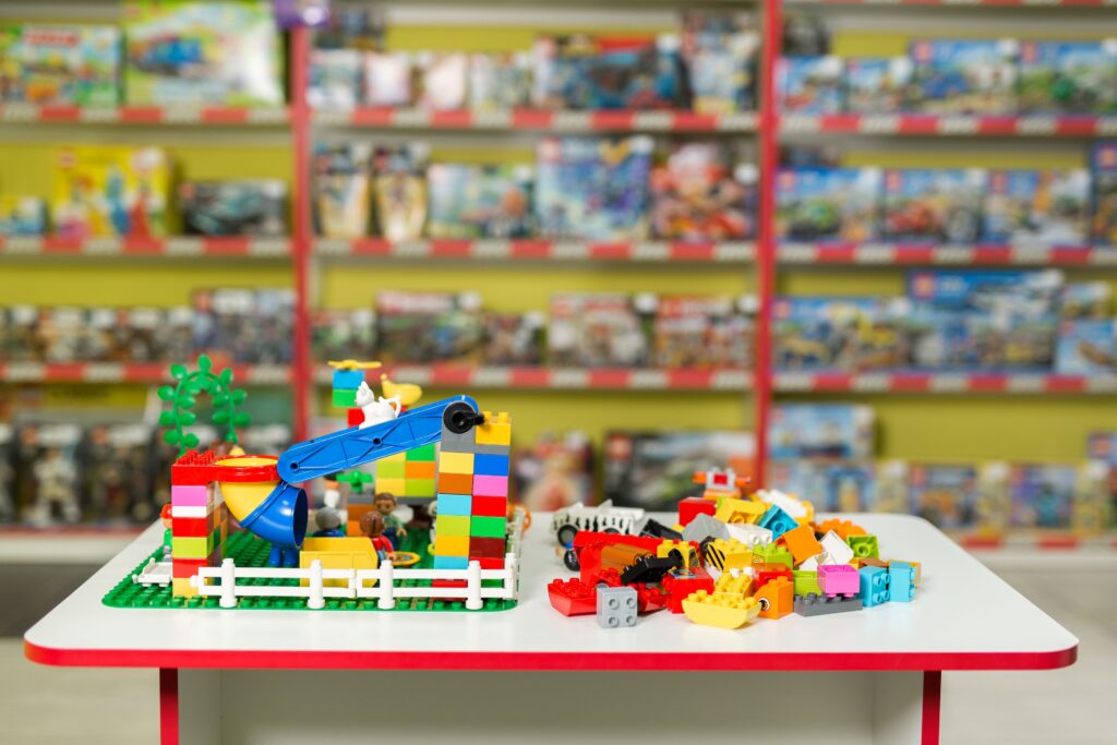 Uma mesa branca exibe uma construção colorida de peças de montar Lego ao lado de uma pilha de blocos soltos, posicionada em uma loja repleta de estantes cheias de brinquedos. A composição da imagem é organizada em dois planos distintos: a mesa em primeiro plano, onde se destaca uma pequena estrutura construída e uma coleção de peças avulsas, e o fundo desfocado, que mostra prateleiras de uma loja de brinquedos. O foco está centralizado nos objetos sobre a mesa, enquanto a profundidade de campo reduzida transforma os produtos nas prateleiras em formas abstratas e coloridas, criando um contraste entre a ordem da criação montada e o estoque comercial atrás. O elemento central é uma estrutura montada com blocos de Lego, caracterizada por pilares de cores primárias como vermelho, amarelo e azul, incluindo uma pequena rampa ou escorregador e uma cerca branca que delimita a área. Dentro dessa estrutura, pequenos bonecos representando figuras humanas estão posicionados como se estivessem interagindo com o ambiente. À direita dessa construção, encontra-se uma pilha de blocos de montar soltos, apresentando uma variedade de formas, tamanhos e cores, sugerindo uma atividade criativa em andamento ou um momento de pausa na brincadeira. A fotografia possui uma estética clara e informativa, típica de registros de ambientes de varejo ou áreas de recreação infantil. A execução técnica prioriza a nitidez dos objetos na mesa, utilizando uma abertura de lente que isola o assunto principal do ambiente circundante. O estilo é direto e funcional, focando na apresentação dos produtos e na atmosfera lúdica do local, com uma iluminação uniforme que elimina sombras fortes e permite que as cores vibrantes das peças de plástico sejam exibidas com clareza. O cenário é o interior de uma loja de brinquedos bem organizada, onde prateleiras com fundos amarelados sustentam inúmeras caixas de jogos e conjuntos. A iluminação ambiente é suave e neutra, típica de interiores comerciais, o que contribui para uma atmosfera convidativa e alegre. A disposição dos elementos cria um ambiente focado no lúdico, onde a criatividade e o entretenimento são destacados tanto pela construção montada sobre a mesa quanto pela vasta oferta de produtos visível no plano de fundo, evocando um sentimento de curiosidade e diversão infantil.