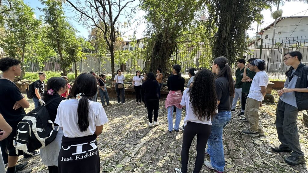 A imagem mostra um grupo de jovens e adultos reunidos em um espaço arborizado, aparentemente participando de uma conversa ou atividade guiada. Algumas pessoas estão em pé, formando um círculo, enquanto outras observam ao redor. O local tem árvores grandes, vegetação, folhas secas no chão e um banco de pedra. Ao fundo, há uma cerca metálica e casas residenciais. A cena é iluminada pelo sol, criando um ambiente tranquilo e ao ar livre.