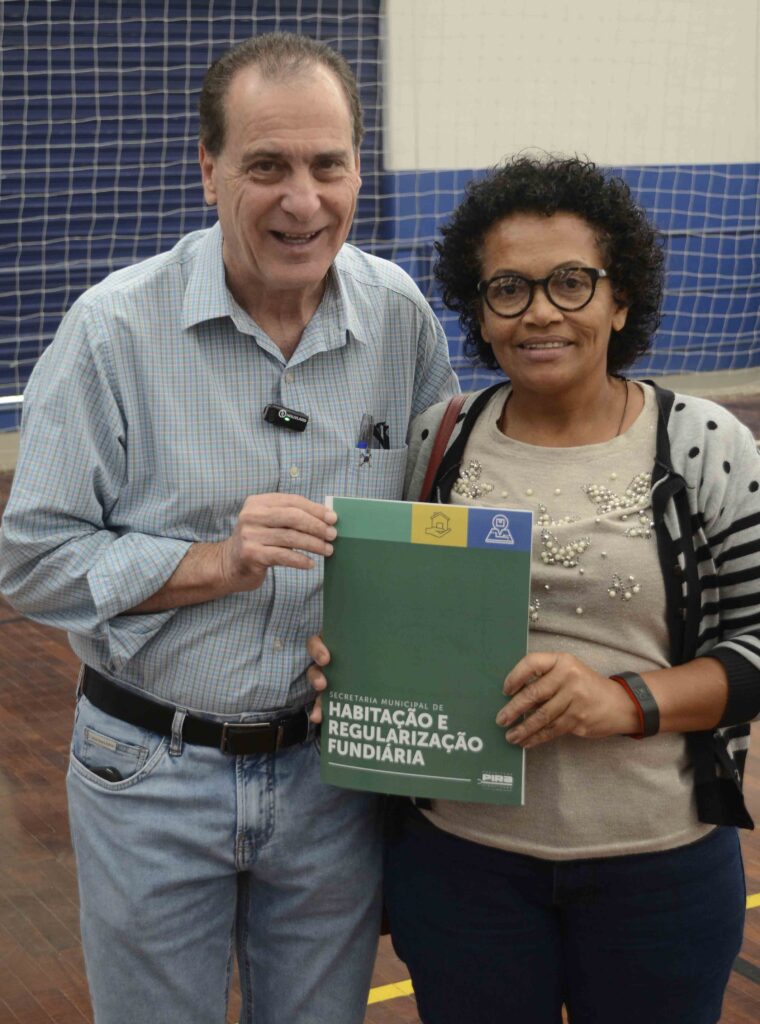 A imagem mostra duas pessoas sorrindo e segurando uma pasta verde da Secretaria Municipal de Habitação e Regularização Fundiária. O homem, de camisa azul clara, e a mulher, de suéter com detalhes de pérolas, parecem participar de uma cerimônia oficial de entrega de documentos. O fundo mostra uma quadra esportiva com rede azul, indicando que o evento ocorre em um espaço comunitário. A cena transmite um momento de conquista e reconhecimento social.