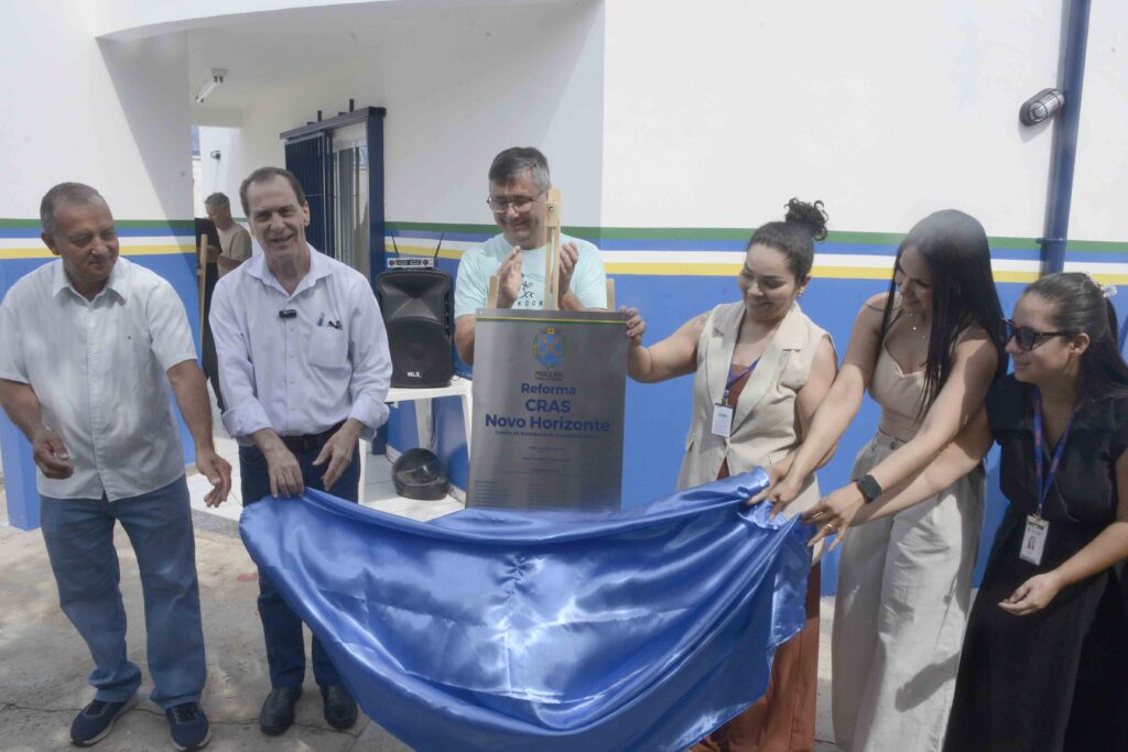 A imagem mostra um grupo de pessoas participando de uma cerimônia de inauguração ou reinauguração de um prédio público. Elas estão revelando uma placa metálica que indica a reforma do CRAS Novo Horizonte, um Centro de Referência de Assistência Social. O grupo, composto por homens e mulheres, parece formado por autoridades e profissionais da área, todos sorridentes e vestidos de forma formal. O ambiente é claro e bem cuidado, com paredes brancas e faixas azul e amarela, transmitindo um clima de celebração institucional.