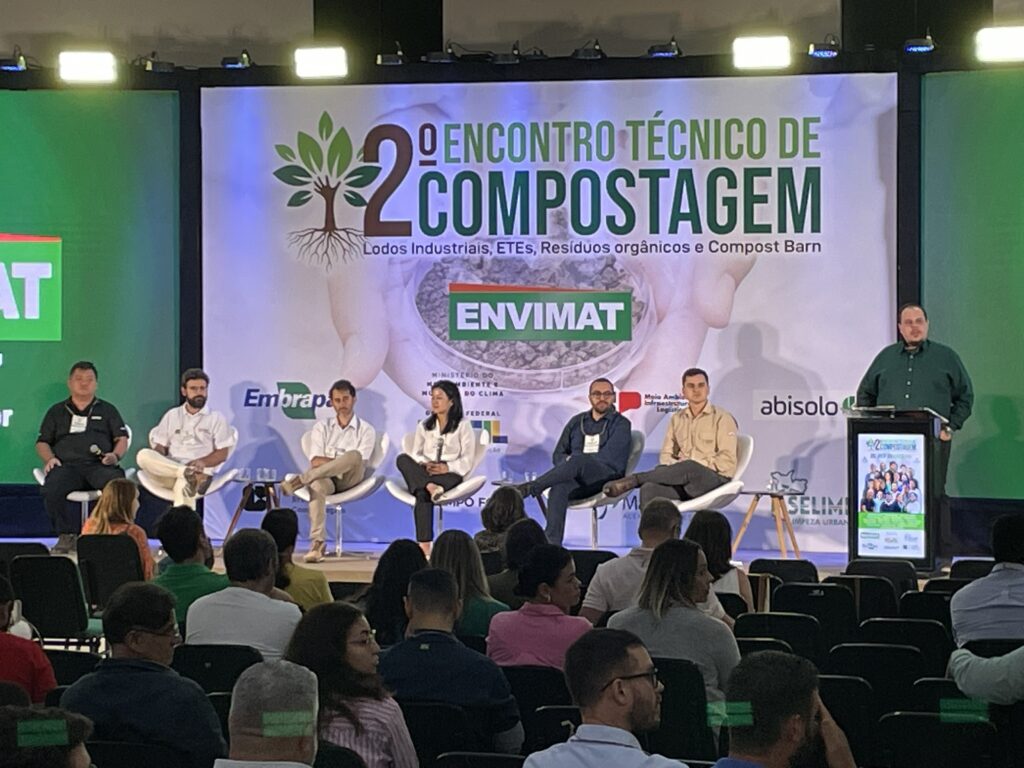 A imagem retrata um ambiente profissional de conferência, onde sete palestrantes estão sentados em um palco diante de uma plateia atenta. Ao fundo, um grande banner anuncia o “2º Encontro Técnico de Compostagem”, destacando temas como lodos industriais, estações de tratamento de esgoto (ETEs), resíduos orgânicos e compost barn. O painel é acompanhado por logotipos de instituições ligadas ao meio ambiente e à agricultura.