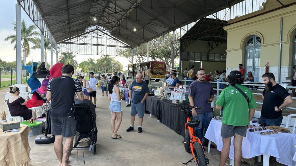 Um grupo de pessoas interage e compra em uma feira ao ar livre, sob uma estrutura coberta.
A cena se passa em uma feira ao ar livre, possivelmente um mercado popular ou feira de artesanato, sob uma grande estrutura coberta com telhado de metal e pilares de sustentação visíveis. A iluminação é natural, com a luz do dia entrando pelas laterais e refletindo nos objetos. Há muitas pessoas presentes, algumas interagindo com barracas de venda, outras conversando e passeando.
No primeiro plano, à esquerda, uma mulher deitada em uma cadeira e vestida com um vestido escuro, com adereços de cabeça coloridos, como chapéus de feiticeiro, em um display próximo. Um homem de costas, vestindo uma camiseta preta com estampa gráfica e shorts cinza, está perto dela, de frente para a câmera. Ao lado deles, há um carrinho de bebê preto.
No centro da imagem, um homem mais velho, vestindo uma camiseta azul escura com a inscrição "California" e shorts cinza, conversa com uma jovem mulher que veste uma top branca e shorts jeans curtos. Atrás deles, há várias barracas com produtos expostos, como artesanato, bijuterias e outros itens. Uma bicicleta elétrica laranja vibrante está estacionada em frente a uma das barracas.
Mais ao fundo, a feira continua com mais barracas e pessoas. Uma van estilizada com a inscrição "Mister Churros Food Bus" está visível, sugerindo opções de comida. Há também edifícios com arquitetura colonial ao fundo, com janelas arqueadas e paredes de cor creme. As cores predominantes na imagem são tons terrosos, azuis, verdes e vermelhos, refletindo tanto as roupas das pessoas quanto os produtos e a ambientação. A atmosfera geral é animada e casual, sugerindo um dia agradável de compras e socialização.