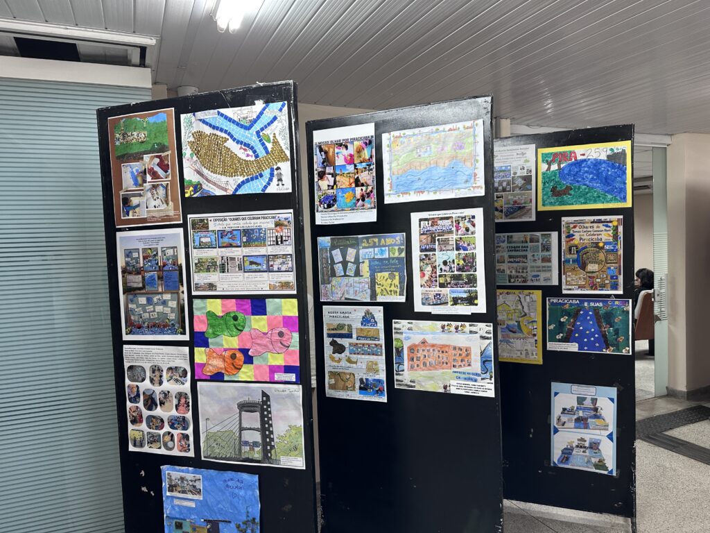 A imagem mostra uma exposição escolar montada em grandes painéis pretos, com desenhos, pinturas, colagens e fotografias sobre a cidade de Piracicaba. É possível ler títulos como “Olhares que celebram Piracicaba”, “Piracicaba 251 anos” e “Piracicaba aos olhos da infância”. Os trabalhos retratam o rio Piracicaba, pontes, paisagens, prédios, bairros, animais, pontos turísticos e memórias da cidade. Há também atividades feitas por crianças, com peixes coloridos, mapas, cenas urbanas e registros de projetos escolares. A exposição está em um espaço interno, sob um teto metálico e bem iluminado.