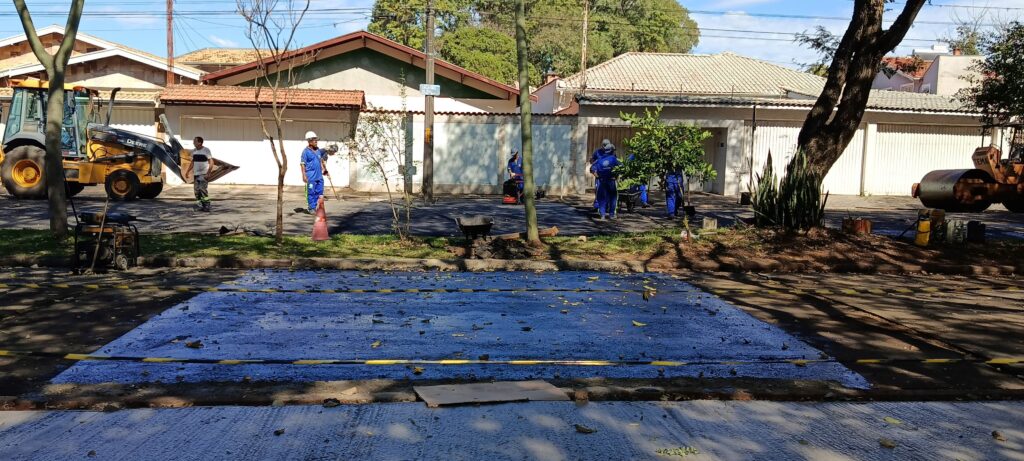 Esta foto mostra uma cena de trabalho de infraestrutura urbana em uma rua residencial. Aqui estão os detalhes: A Ação: Há uma equipe de trabalhadores vestindo uniformes azuis e capacetes realizando um serviço no pavimento. Alguns deles estão espalhados pela via, e um dos funcionários está usando uma máquina de compactação de solo (o "sapinho"). O Destaque (Pintura Azul): Em primeiro plano, no chão, há um grande retângulo pintado de azul vibrante que parece ser uma marcação, possivelmente para uma faixa de pedestres ou sinalização especial. Ela está delimitada por faixas zebradas de sinalização amarela e preta. Equipamento: No lado esquerdo, vemos uma retroescavadeira da marca "John Deere" amarela, e no lado direito, parcialmente visível sob uma árvore, está um rolo compactador. Ambiente: É um dia ensolarado em um bairro residencial. Ao fundo, vemos casas com muros brancos e portões de garagem, e a rua é bem arborizada, projetando sombras sobre o solo. Há também uma placa de sinalização de rua indicando "Cruzeiro do Sul".