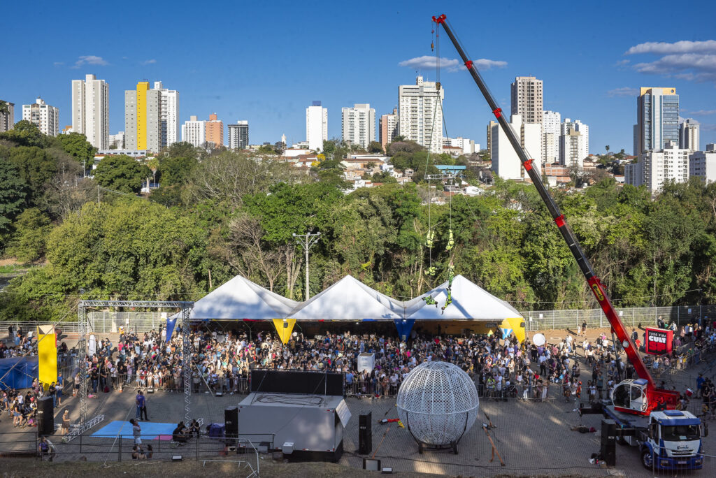 A imagem mostra um grande evento ao ar livre em uma área urbana, com muitas pessoas reunidas sob tendas brancas e coloridas. Um guindaste vermelho ergue dois artistas suspensos no ar, vestidos com roupas verdes, realizando uma performance acrobática. No chão, há uma estrutura metálica esférica — típica de apresentações com motocicletas — e equipamentos de palco. Ao fundo, vê-se uma cidade com prédios altos e modernos cercados por árvores, sob um céu azul claro. A cena transmite energia e entusiasmo, típica de um festival de circo ou espetáculo público.