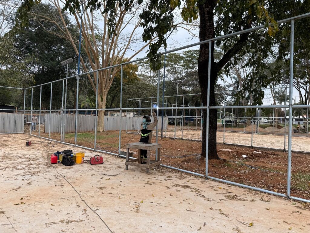 A imagem mostra um trabalhador soldando ou instalando uma estrutura metálica cercada, provavelmente em uma quadra ou área esportiva de um parque. Ele está sobre uma pequena plataforma de madeira e usa equipamentos de proteção, incluindo capacete, máscara e roupa refletiva. No chão, há ferramentas, cabos, baldes e recipientes de trabalho. Ao redor, aparecem árvores grandes, postes de iluminação, áreas com areia e terra, além de outros trabalhadores e estruturas em construção.