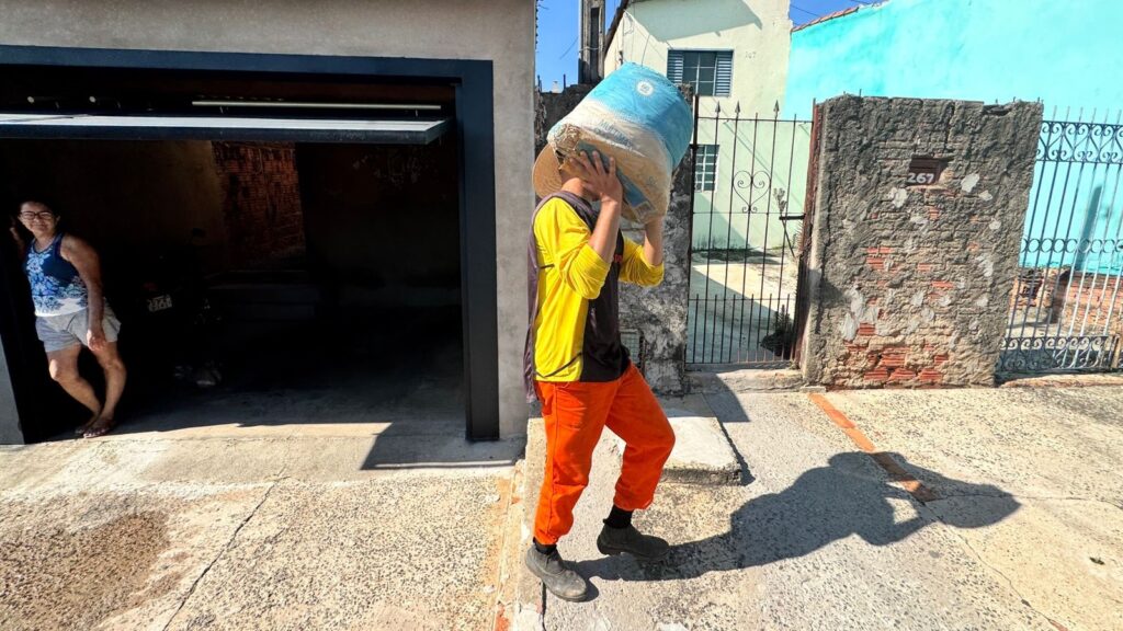 Pessoa carregando um saco grande sobre os ombros, andando na calçada em frente a uma casa. Pessoa: corpo jovem/adulto (rosto coberto pelo saco, não identificável). Usa camiseta amarela com mangas compridas, colete escuro por cima, calça laranja e botas de trabalho pretas. Está com as mãos segurando o saco apoiado na cabeça/ombros. Carga: saco grande, azul e bege, aparenta ser de cimento, ração ou material a granel (apenas aparência visual, sem confirmar conteúdo). Cena e postura: a pessoa avança com uma perna à frente; sombra longa projetada no chão indicando sol forte e horário de luz alta. Ambiente imediato: entrada de garagem à esquerda (porta parcialmente aberta) com uma mulher observando da sombra; muro e portão de ferro à direita, com placa de número 267 na coluna de cimento/tiijolo. Casa ao fundo pintada em tons claros e um muro azul-claro mais adiante. Piso e iluminação: calçada de concreto com pequenas fissuras e manchas; iluminação direta e sombras nítidas, dia ensolarado. Atmosfera geral: cena cotidiana de trabalho/entrega em área residencial; contraste claro/escuro entre a sombra da garagem e a rua iluminada. Descrevi apenas o que é visível na imagem, sem identificar pessoas.