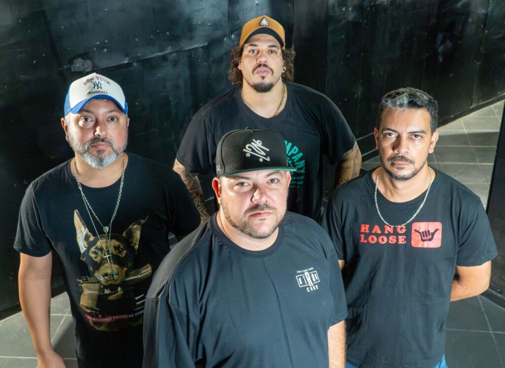 A imagem retrata um grupo de quatro homens, todos vestidos com camisetas pretas e usando bonés. Eles parecem estar em um ambiente com fundo escuro e texturizado, possivelmente uma parede metálica ou industrial, com alguns reflexos de luz. Da esquerda para a direita, o primeiro homem, que está mais à frente e ligeiramente à esquerda, usa um boné branco e azul com o logo "New York Yankees". Ele tem uma barba grisalha e usa uma corrente prata com um pingente em forma de cruz sobre uma camiseta preta com a estampa de um cachorro que morde uma bandoleira. Há uma inscrição em português abaixo da imagem do cachorro, parcialmente visível. No centro, um pouco mais atrás e ocupando a parte central inferior da imagem, está um homem com um boné preto com um logo branco estilizado. Ele tem uma barba curta e um olhar direto para a câmera. Sua camiseta preta tem um pequeno logo quadrado branco com letras verdes no lado direito do peito. Atrás e no centro, ligeiramente mais alto, está outro homem com um boné marrom com um pequeno símbolo dourado. Ele tem cabelos cacheados escuros e uma barba curta. Sua camiseta preta exibe parte de um logo verde com o texto "JAPAN" visível. Ele também usa uma corrente fina dourada. À direita, e mais à frente, está um homem com uma camiseta preta com a estampa vermelha "HANG LOOSE" acompanhada do símbolo do "shaka" (sinal de mão do surfista). Ele tem cabelos escuros curtos e um bigode fino. Usa uma corrente prata. A composição é centrada no grupo, com os homens posicionados de forma a preencher o quadro. A iluminação parece direcionada, destacando os rostos e as estampas das camisetas, enquanto o fundo permanece mais escuro e desfocado, criando um contraste que enfatiza os sujeitos. As cores predominantes são o preto das roupas e do fundo, com toques de branco, azul, marrom, dourado, verde e vermelho nas estampas e bonés. As expressões dos homens são sérias e diretas para a câmera, transmitindo uma pose deliberada e confiante. Há detalhes visíveis como tatuagens em alguns dos homens.