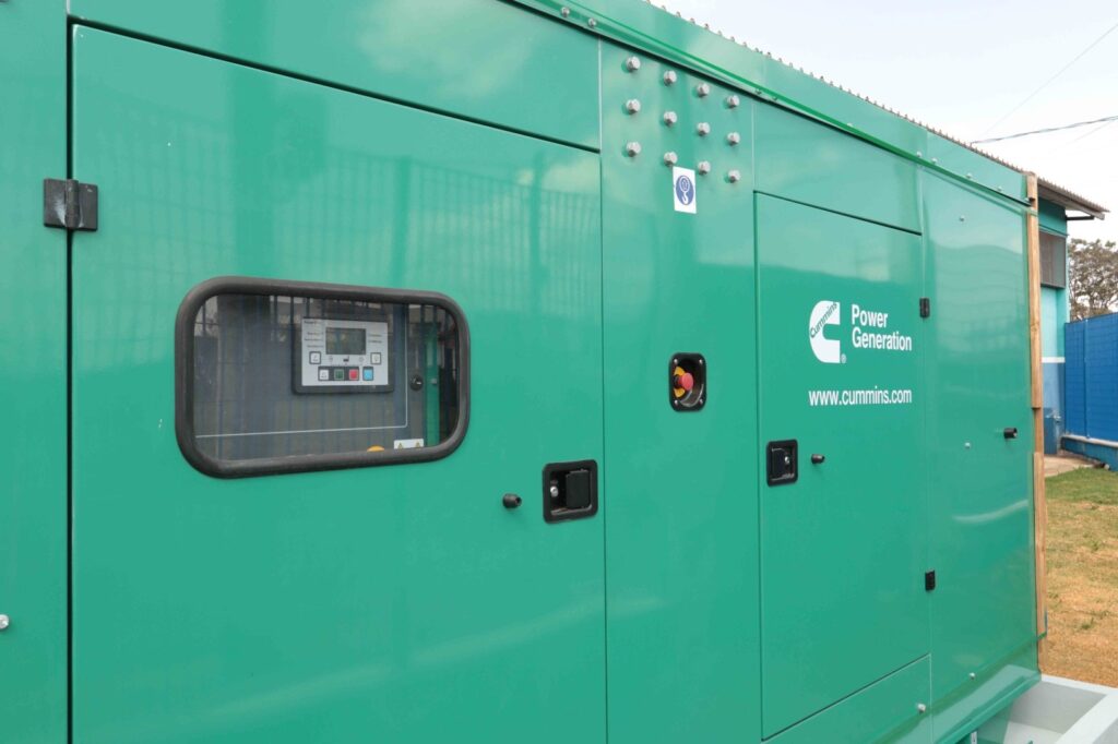 A imagem mostra um gerador elétrico industrial da marca Cummins Power Generation, revestido por uma carcaça verde. Na lateral, há uma janela que permite visualizar o painel de controle, além de portas de acesso, dobradiças, parafusos e um botão vermelho de emergência. Também aparecem o logotipo da Cummins e o endereço www.cummins.com. O equipamento está instalado ao lado de um prédio, próximo a uma cerca azul e a uma área com gramado seco.