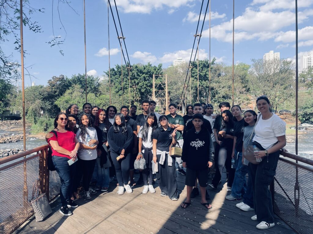 A foto mostra um grupo grande de pessoas reunido em uma ponte suspensa de madeira sobre um rio. O grupo posa para a câmera, com adultos e jovens usando roupas casuais. Ao redor há árvores, vegetação, pedras e água corrente; ao fundo, aparecem prédios altos. O céu está azul, com algumas nuvens, e a cena é iluminada pelo sol.