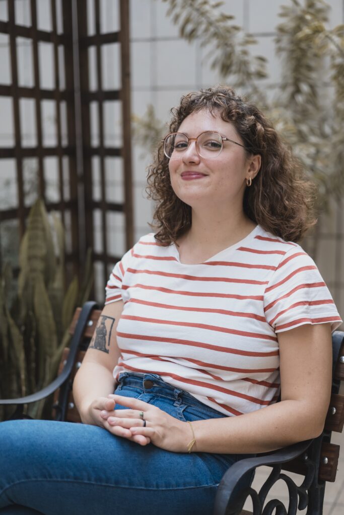 Esta foto mostra uma jovem sorrindo, sentada de forma tranquila em um banco de jardim. Aqui estão os detalhes: A Pessoa: Ela tem cabelos cacheados e castanhos que caem suavemente sobre os ombros. Usa óculos de grau com armação redonda em tom metálico e argolas pequenas na orelha. Sua expressão é serena e simpática. Vestimenta: Ela veste uma camiseta de listras horizontais brancas e vermelhas e calças jeans azul-claro. É possível notar uma tatuagem no braço direito, que parece representar uma figura ilustrativa. Pose: Ela está sentada com as mãos entrelaçadas sobre o colo, transmitindo uma postura relaxada e confortável. Ambiente: O cenário parece ser uma área externa ou uma varanda. Atrás dela, há uma treliça de madeira e algumas plantas (como uma espada-de-são-jorge), o que dá um toque natural e acolhedor à cena. A iluminação é suave e natural.