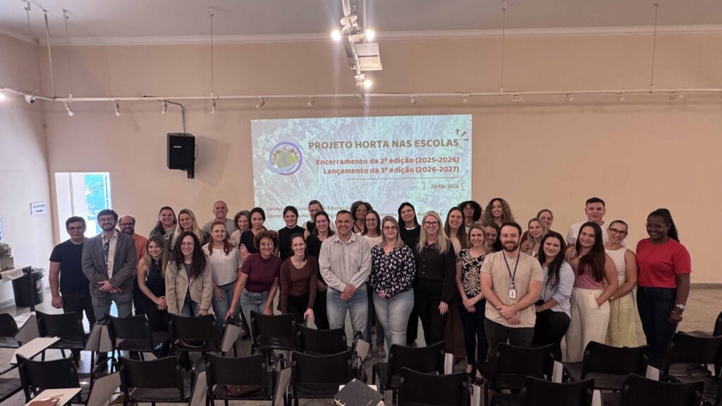 A imagem mostra um grupo numeroso de adultos reunido em uma sala, posando para uma foto. Algumas pessoas estão sentadas e outras permanecem em pé, todas voltadas para a câmera. Ao fundo, há uma tela de projeção com o título “Projeto Horta nas Escolas”, indicando o encerramento da 2ª edição (2025–2026) e o lançamento da 3ª edição (2026–2027), com a data 24/08/2026. O ambiente possui cadeiras, equipamentos de som, iluminação de trilhos e uma placa indicando o banheiro.