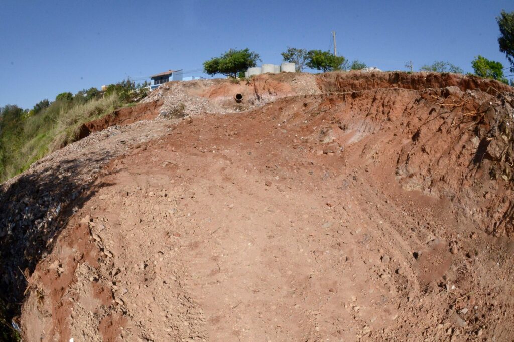 Na foto, vemos um trecho de encosta erodida com solo exposto em tons avermelhados e presença de pedras soltas. No alto da área, sobressai um tubo de drenagem, sugerindo que o escoamento de água contribuiu para o desgaste do terreno. Atrás da erosão, aparecem árvores, vegetação e uma construção, além de objetos cilíndricos de concreto que lembram reservatórios ou materiais de obra. O céu azul e claro reforça o registro diurno, enquanto a cena evidencia os impactos da degradação do solo possivelmente ligada a falhas de drenagem ou atividades de construção próximas à borda da encosta.