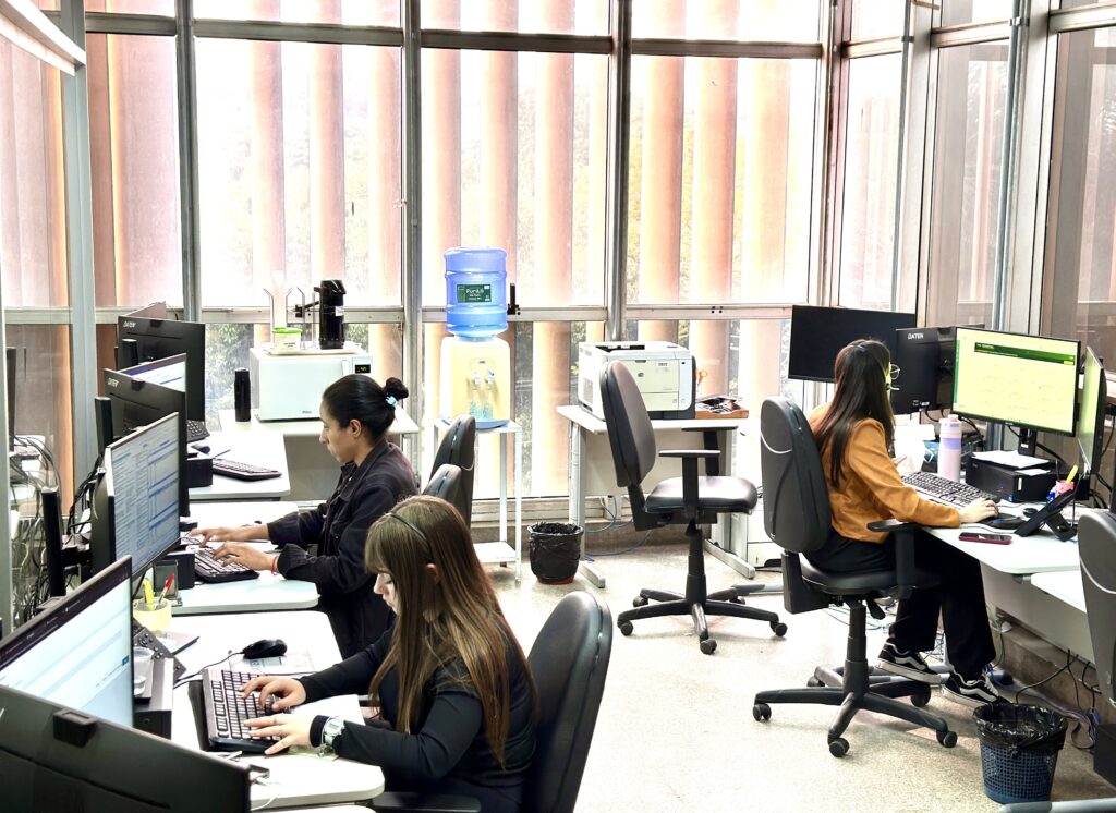 A imagem mostra um ambiente de escritório funcional, com três pessoas trabalhando em suas estações de computador. Aqui estão os pontos principais: Configuração: O espaço é organizado com mesas individuais, cada uma com monitor, teclado e mouse. O ambiente tem grandes janelas que ocupam toda a parede lateral, permitindo bastante luz natural. Equipe: Duas pessoas estão sentadas à esquerda, concentradas em suas telas. Uma pessoa está sentada à direita, também trabalhando em um computador com monitor grande. Equipamentos: Além dos computadores, há um bebedouro de galão ao fundo, uma impressora e alguns itens de escritório espalhados, como garrafas de água e materiais de papelaria. Ambiente: O espaço parece ser um escritório administrativo ou de suporte, com uma atmosfera de trabalho focada. O piso é de granilite claro.