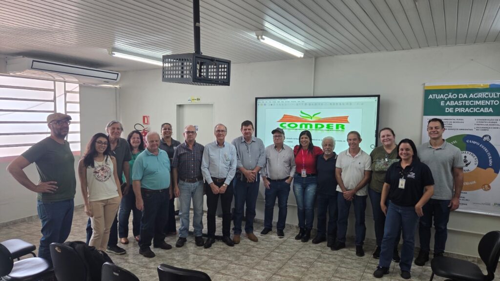 Vejo um grupo de 15 pessoas posando em uma sala de reunião/treinamento: Disposição: todos em pé em linha única à frente da parede, olhando para a câmera e, em sua maioria, sorrindo. Idades e gênero: mistura de homens e mulheres de idades variadas (adultos jovens a idosos). Roupas: traje casual a social-casual — camisas pólo, camisas de botão, blusas, jeans e calças; duas pessoas usam crachá/identificação visível. Ambiente: sala com piso de cerâmica, teto com forro branco e luminárias fluorescentes; ar-condicionado de parede à esquerda. Equipamentos e materiais: ao fundo, um display/tela grande mostrando o logotipo “COMDER”; à direita, um banner vertical com o título “ATUAÇÃO DA AGRICULTURA E ABASTECIMENTO DE PIRACICABA” e gráficos informativos; também há um extintor de incêndio encostado na parede. Mobiliário: cadeiras pretas empilhadas ou deslocadas em primeiro plano e laterais. Iluminação: luz natural entrando pela janela com venezianas à esquerda e iluminação interna clara, cena bem visível. Atmosfera: parece ser um encontro formal/institucional — reunião, apresentação ou workshop ligado a agricultura/abastecimento — com tom cordial e colaborativo.