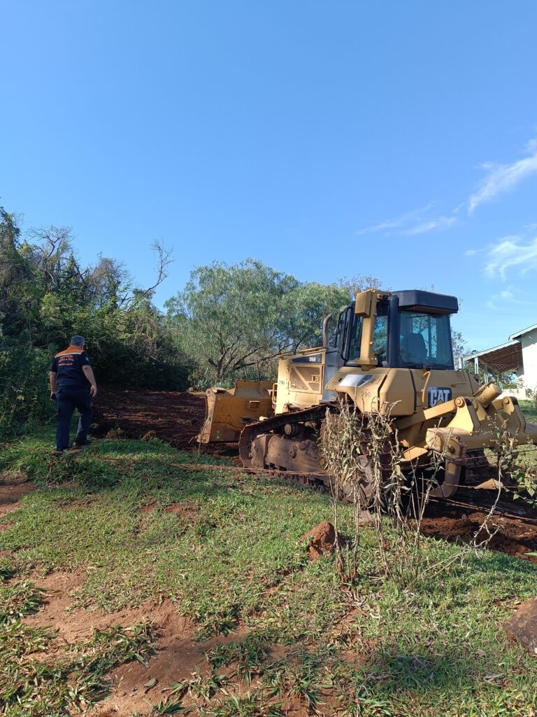 Nesta imagem, vemos uma cena de trabalho ao ar livre em um dia ensolarado. Principal: Um trator de esteira grande, amarelo, da marca CAT (modelo D6N), está parado sobre um terreno de terra batida que parece ter sido recentemente remexido ou preparado. Pessoa: À esquerda, um homem está caminhando em direção à mata lateral. Ele veste um uniforme com a inscrição "Defesa Civil" nas costas. Ambiente: O terreno é uma mistura de terra exposta (onde o trator está) e vegetação rasteira. Ao fundo, há uma área arborizada e, à direita, parte de uma estrutura ou construção coberta. O céu está limpo e azul.