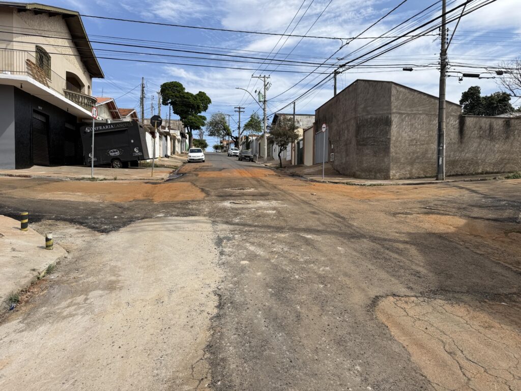 Nesta imagem vemos um cruzamento em um bairro residencial, onde o asfalto apresenta remendos e irregularidades que revelam reparos recentes ou desgaste do tempo. As casas e pequenos comércios se alinham pelas laterais, incluindo uma fachada preta com o letreiro “CONFRARIA LANCHES”. Um carro branco aparece estacionado mais adiante, enquanto postes e fios elétricos atravessam o cenário. Perto da esquina, uma placa de trânsito indica a proibição de virar à esquerda.
