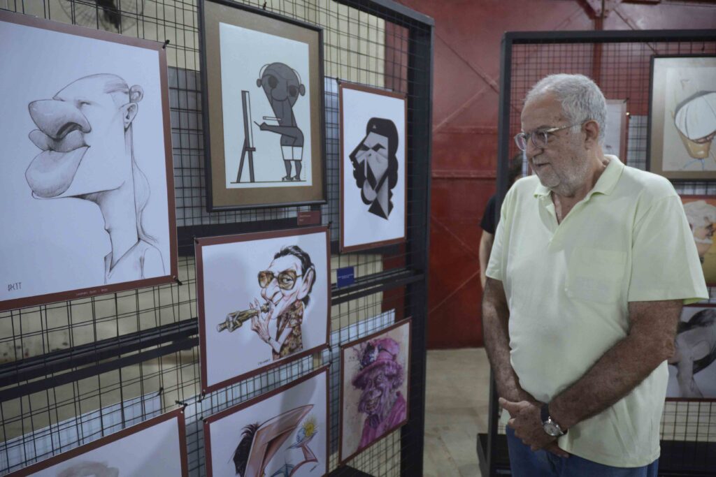 A imagem mostra uma exposição de desenhos e pinturas pendurados em uma grade metálica. Em primeiro plano, há dois retratos de pessoas idosas: à esquerda, um homem de cabelos brancos e óculos; à direita, uma mulher de cabelos grisalhos, também usando óculos. Ao fundo, aparecem outros trabalhos artísticos, incluindo ilustrações coloridas de figuras humanas e flores. No canto inferior esquerdo, vê-se uma pequena anotação manuscrita. A cena parece fazer parte de uma mostra de arte ou atividade cultural.